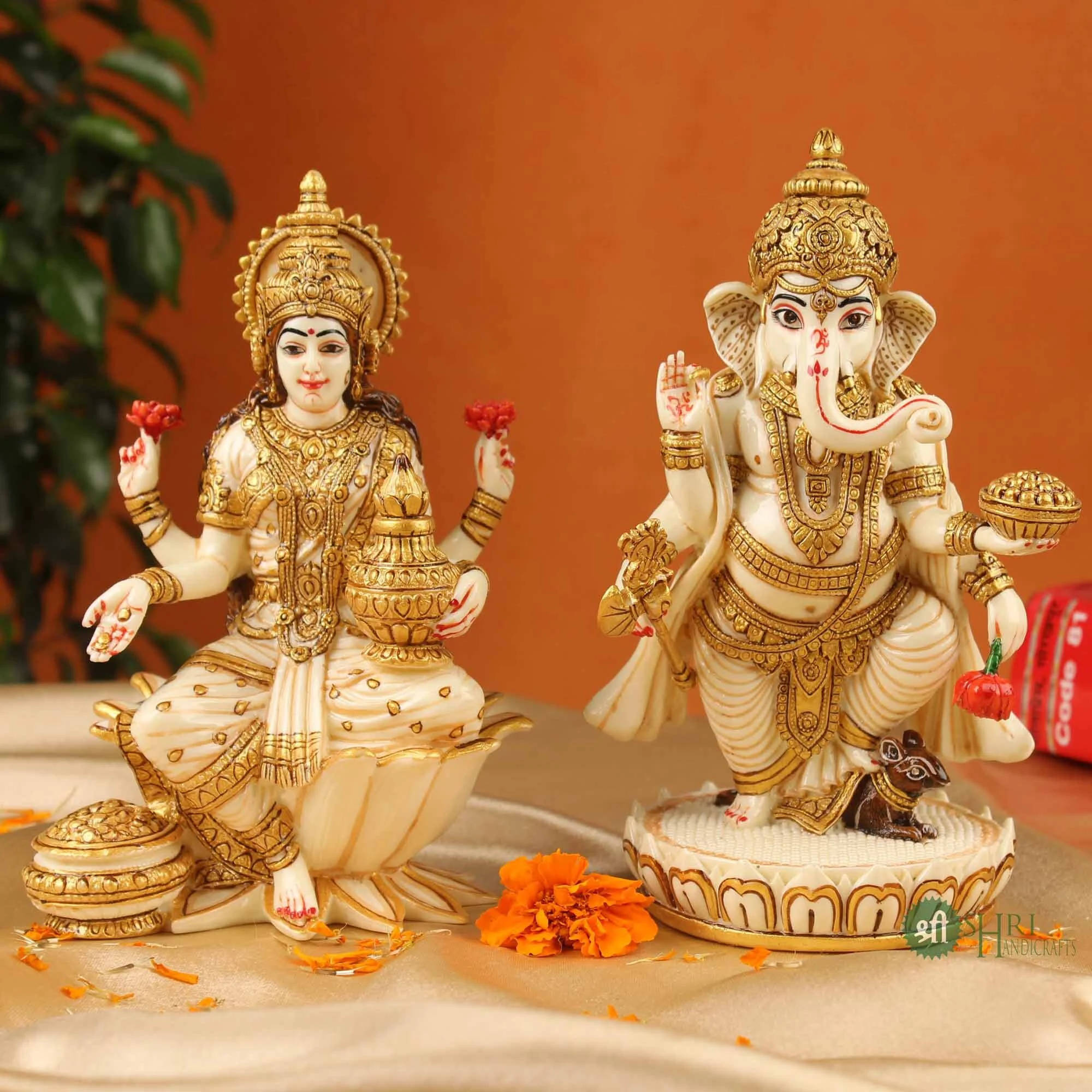 Lakshmi 桌布 壁紙 — 兩尊精雕細琢的印度教神像，象頭神伽內什和吉祥天女拉克什米，飾以金和象牙，優雅地坐在緞面之上，背景有萬壽菊和模糊的綠色植物。