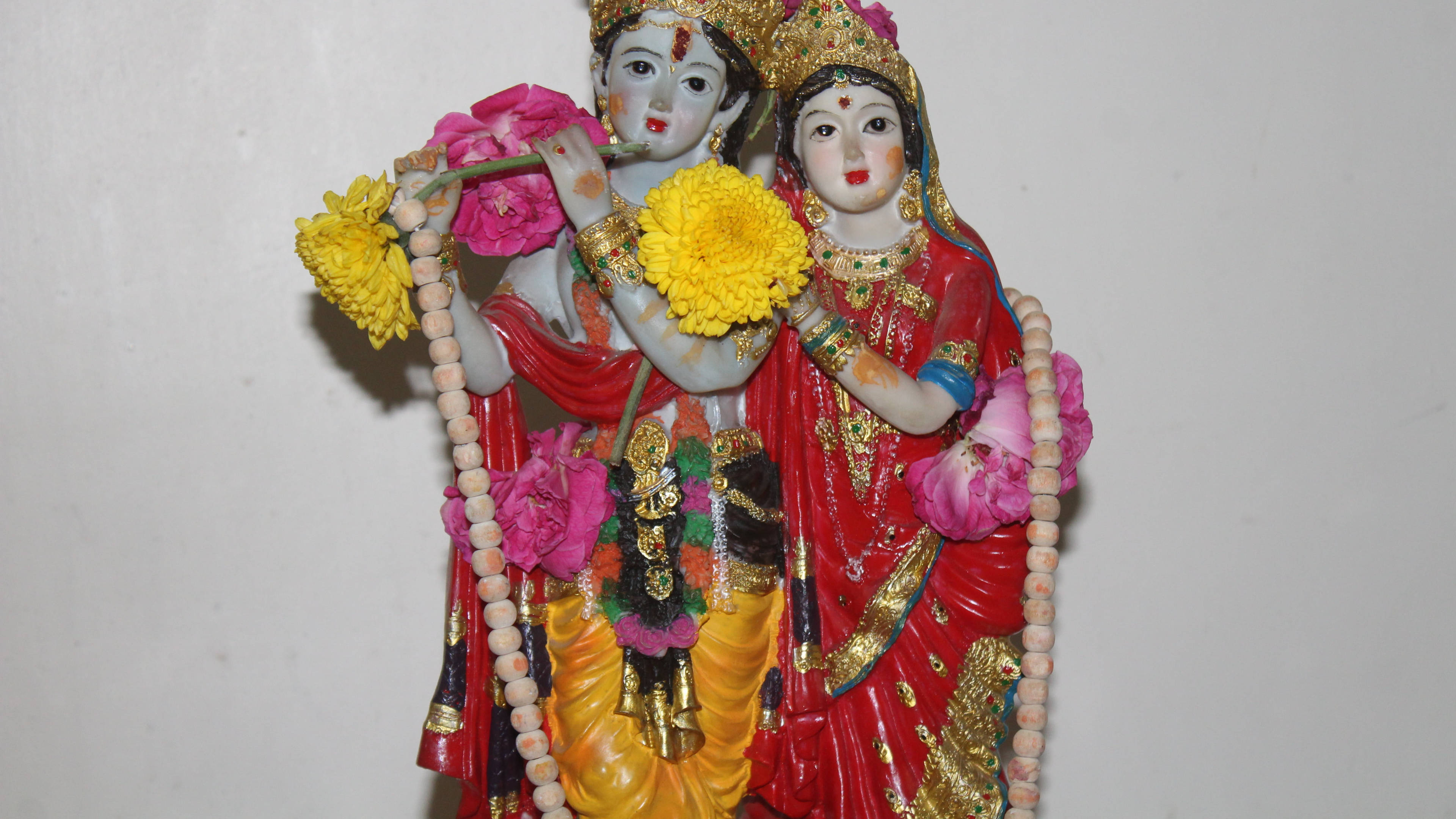 krishna ji 桌布 壁紙 — 兩尊裝飾華麗的印度教神像，身穿鮮豔的紅色、金色和粉紅色服飾，手持鮮花和一支笛子，背景是純白色的牆壁。