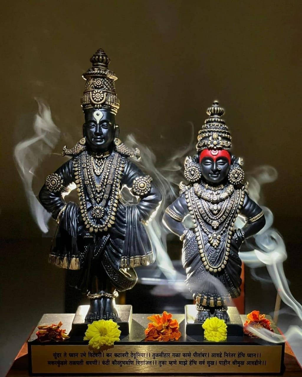 Indian 桌布 壁紙 — 兩尊裝飾華麗的黑色神像，飾有金色珠寶和紅色頭帶，站在一個帶有萬壽菊和裊裊香煙的基座上。