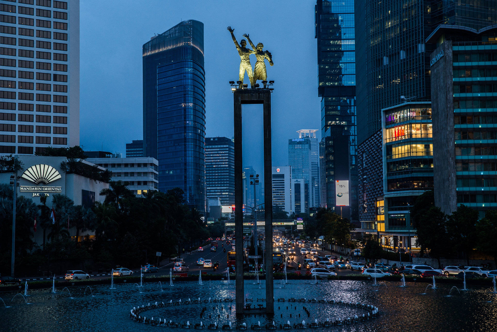 Jakarta 桌布 壁紙 — 兩尊金色雕像矗立在高高的基座上，位於噴泉中，周圍是黃昏時分高聳的摩天大樓。