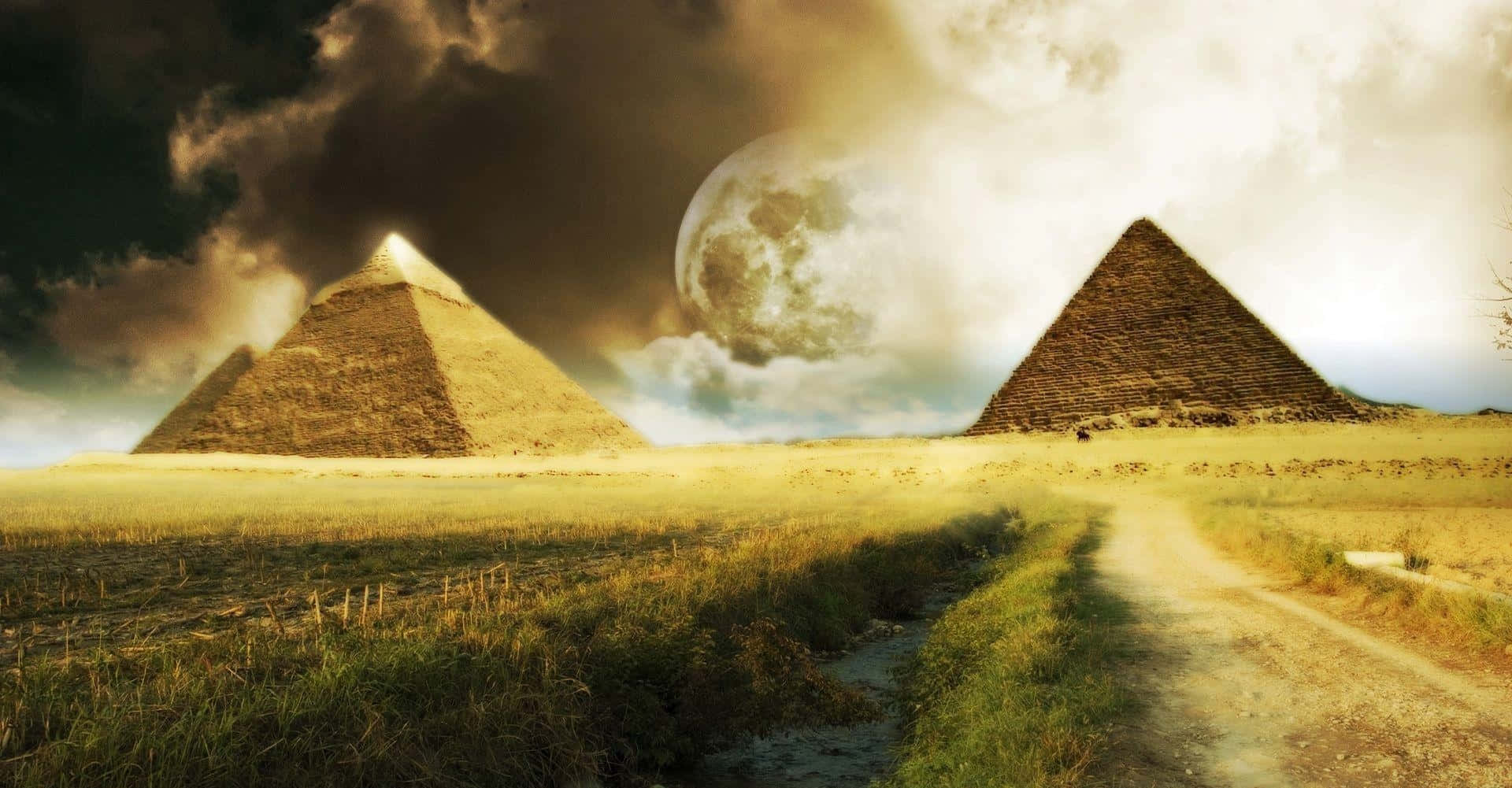 Dark Surrealism 桌布 壁紙 — 兩座古老金字塔矗立在戲劇性的天空下，一輪巨大的月亮將金色光芒灑在一條蜿蜒的泥土小徑上。