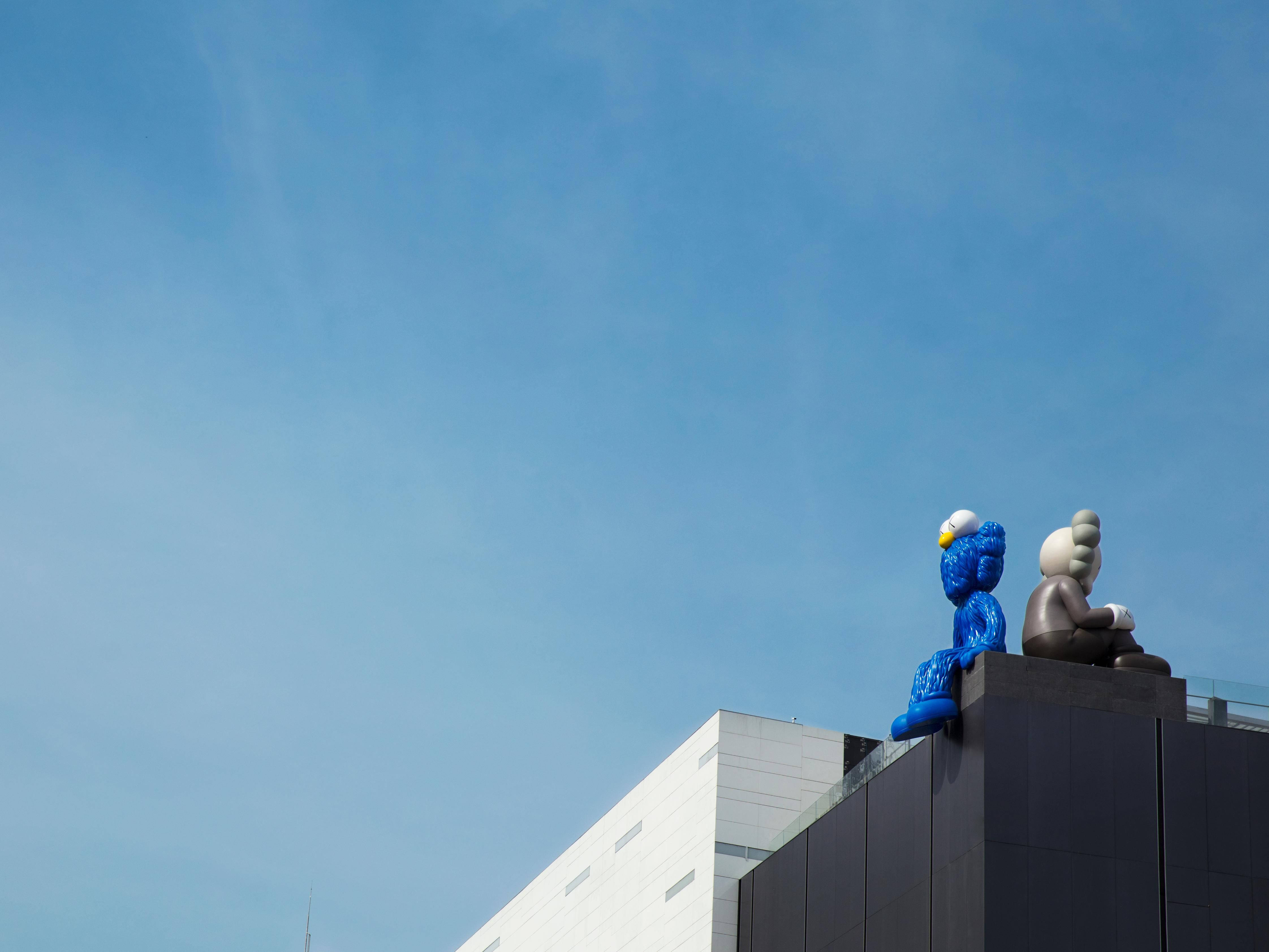 KAWS 桌布 壁紙 — 兩座大型風格化雕塑坐落在現代建築頂部，襯著清澈的藍天。