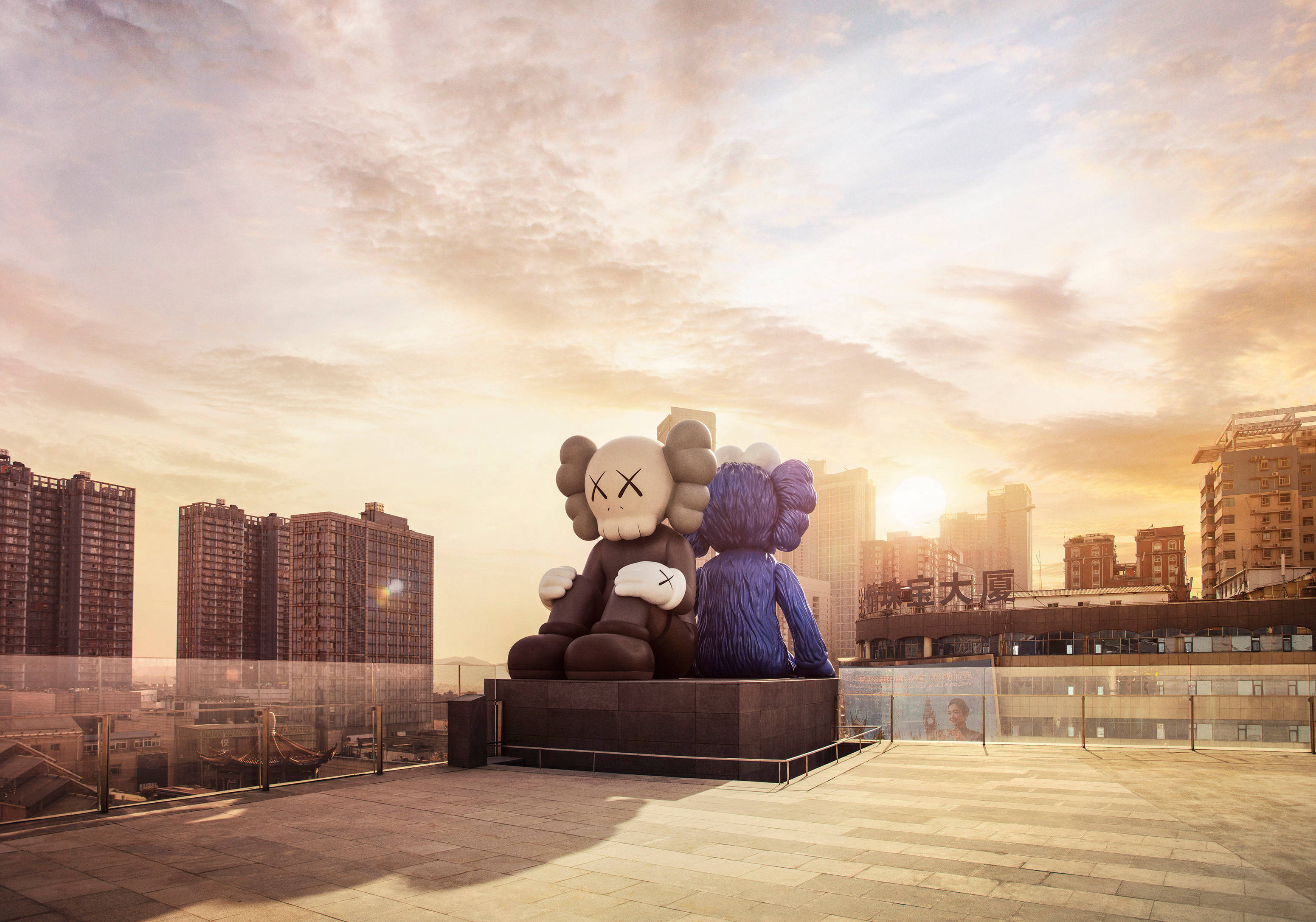 KAWS 桌布 壁紙 — 兩座巨大的KAWS雕塑坐在屋頂上，俯瞰沐浴在金色夕陽光芒中的城市景觀。