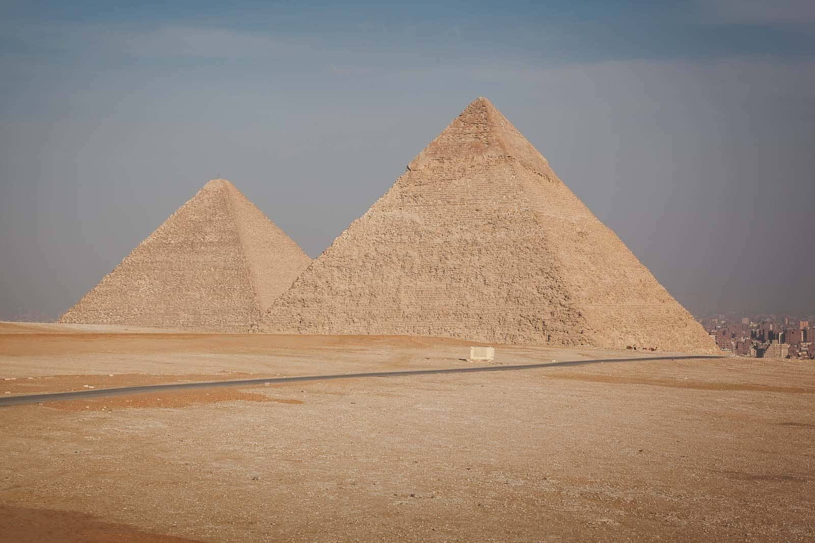 Ancient Egypt 桌布 壁紙 — 兩座巨大的古老金字塔從廣闊的沙漠中升起，在朦朧的藍天下，地平線上有遙遠的城市景觀。