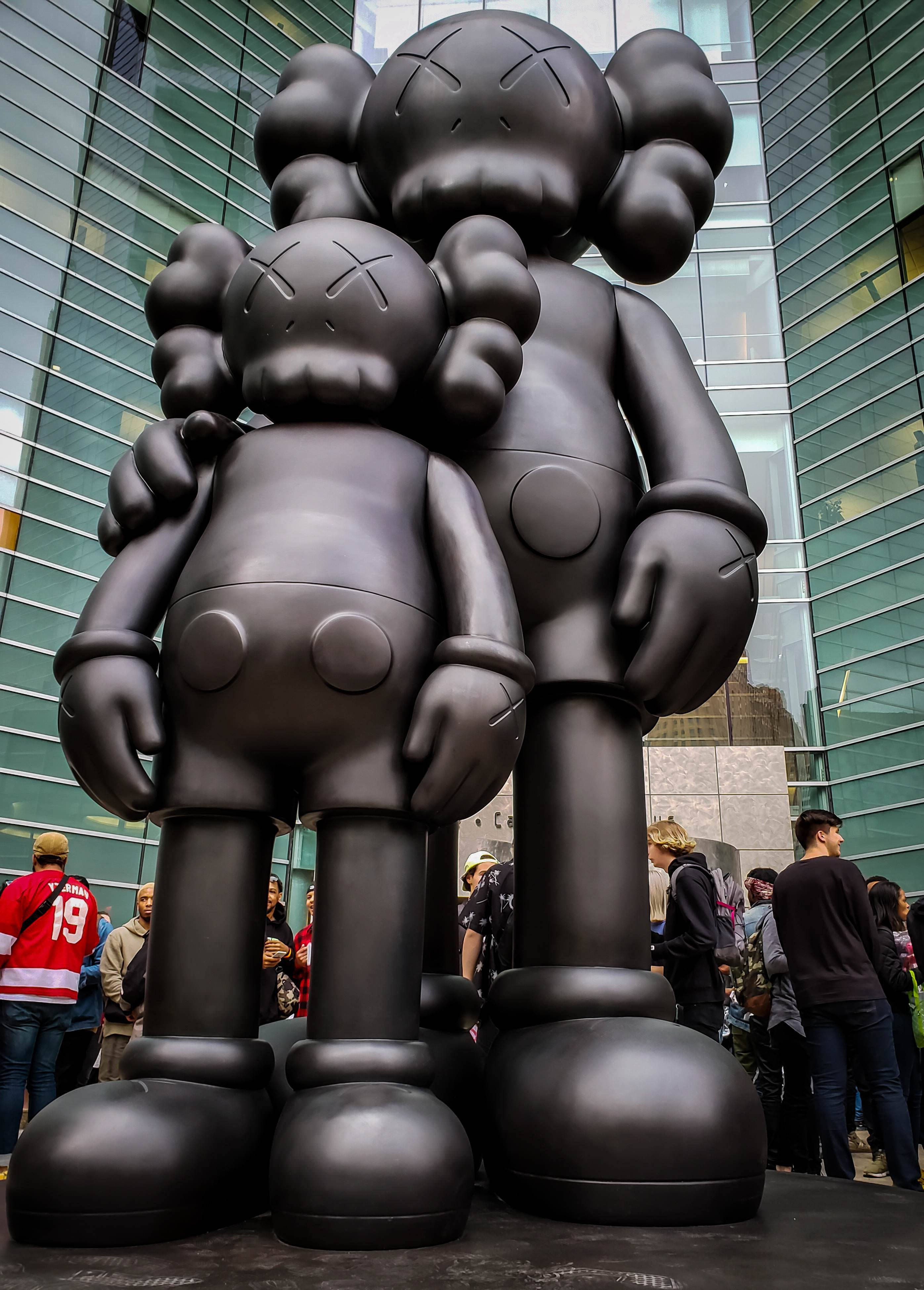 KAWS 桌布 壁紙 — 兩座巨大的啞光黑色KAWS雕塑在玻璃摩天大樓前擁抱，周圍聚集了圍觀者。