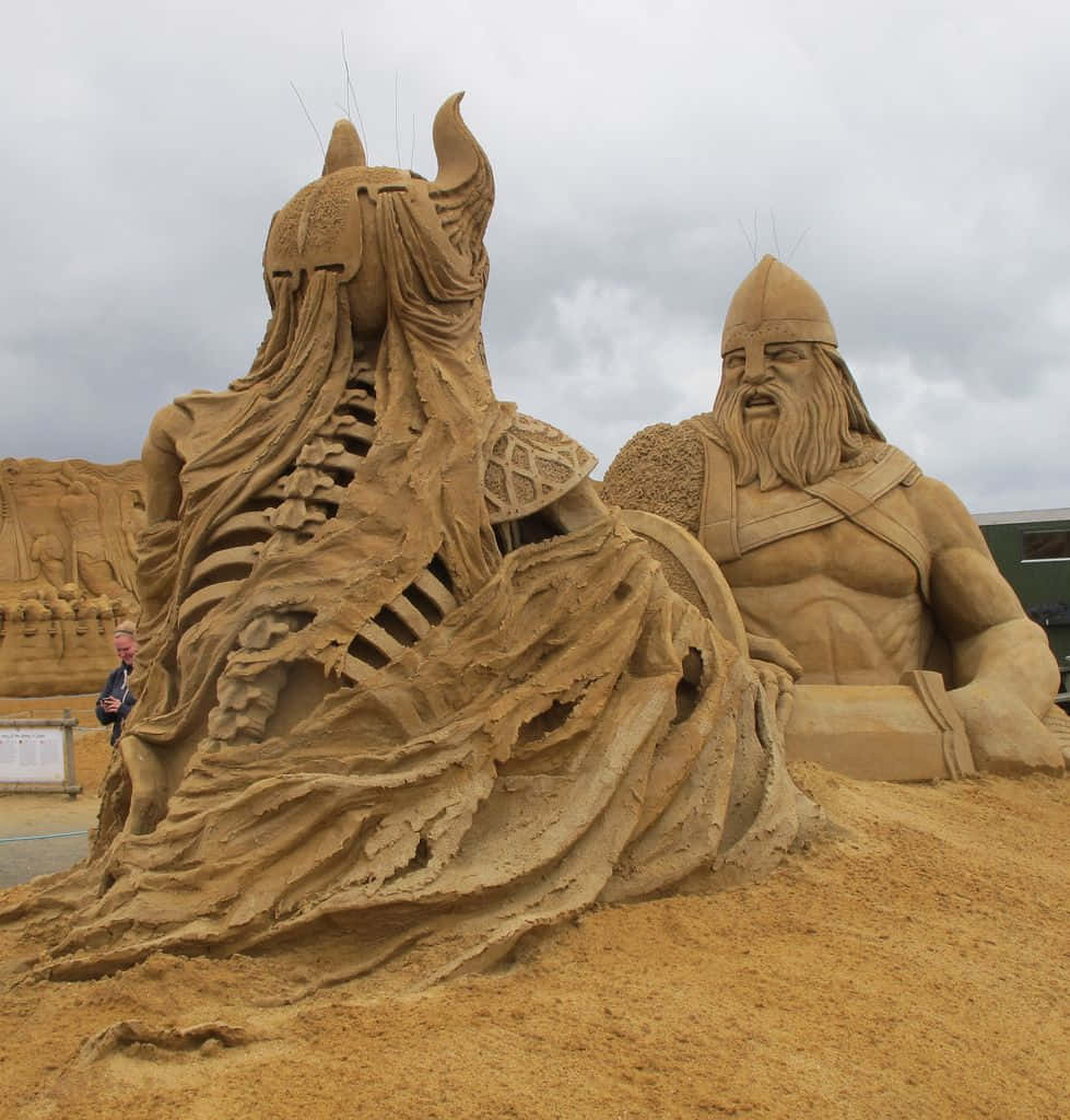 Sand Art 桌布 壁紙 — 兩座巨大的沙雕描繪了一個有角的戰士和一個鬍鬚維京人在陰天下的戲劇性戶外展示。