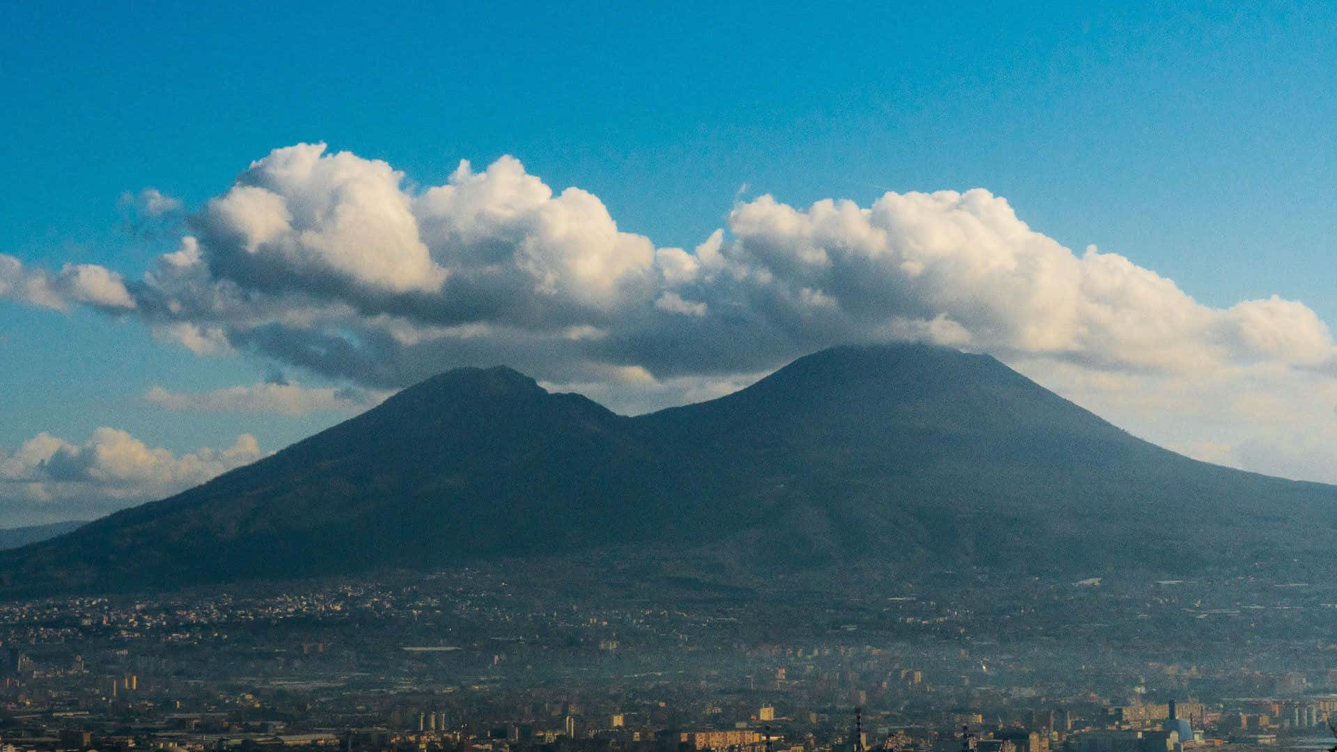 Volcano 桌布 壁紙 — 兩座深色火山聳立於一座廣闊城市之上，天空湛藍，白雲蓬鬆。