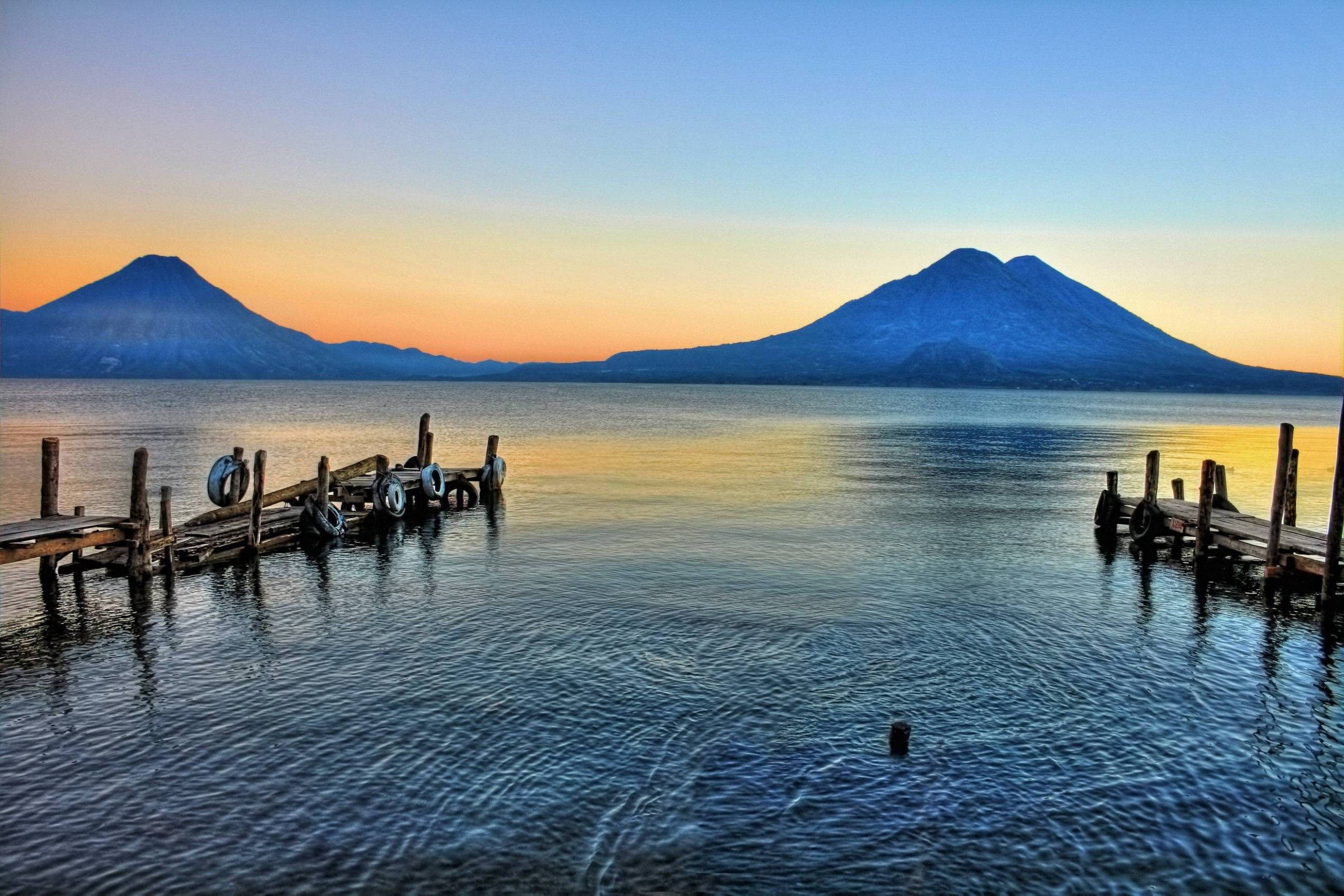 Guatemala 桌布 壁紙 — 兩座火山在日落時分雄偉地聳立在平靜的湖泊後方，木製碼頭延伸至水中。