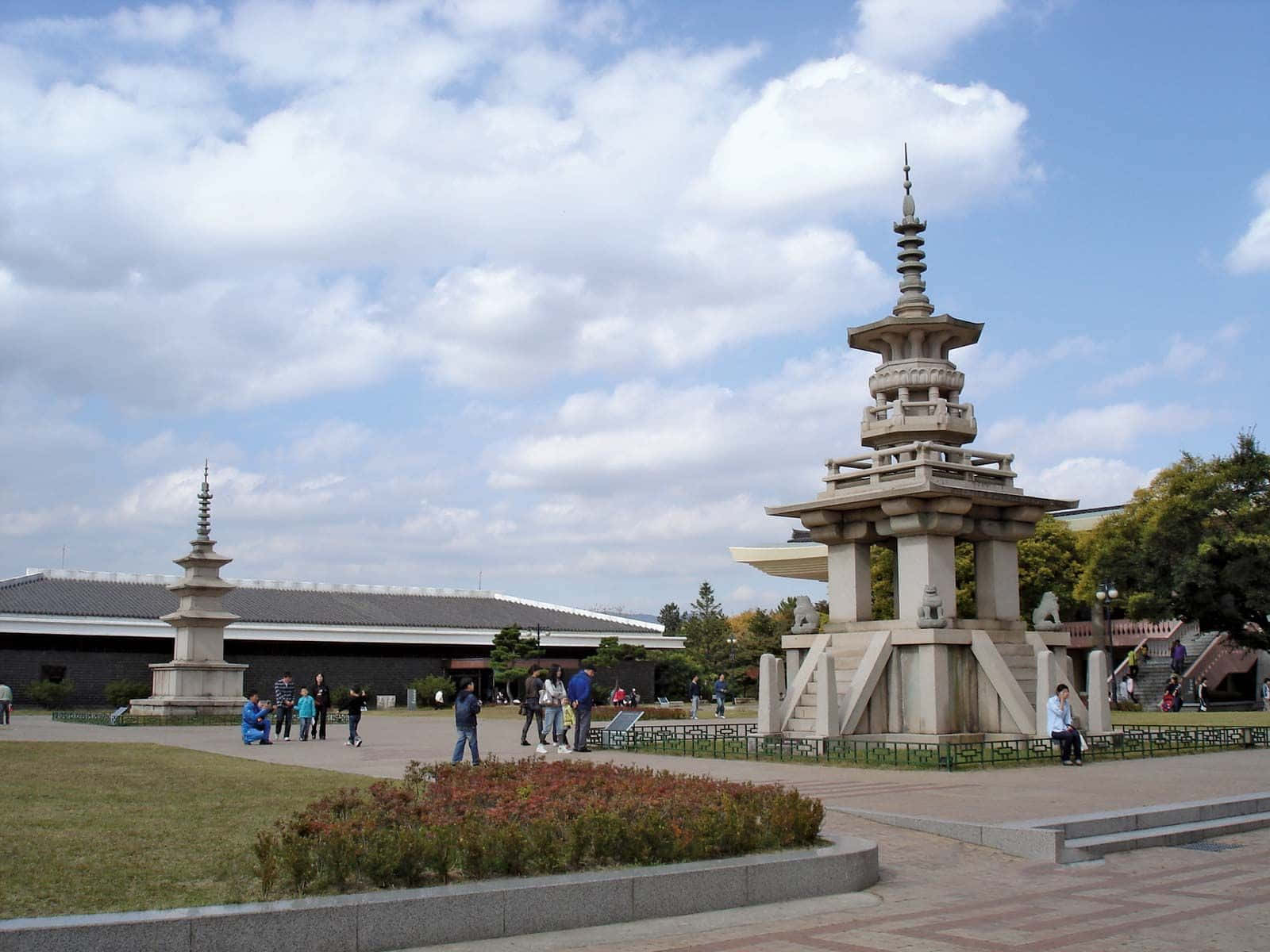 南韓 桌布 壁紙 — 兩座石塔在公園中高高聳立，天空局部多雲，有遊客在漫步。