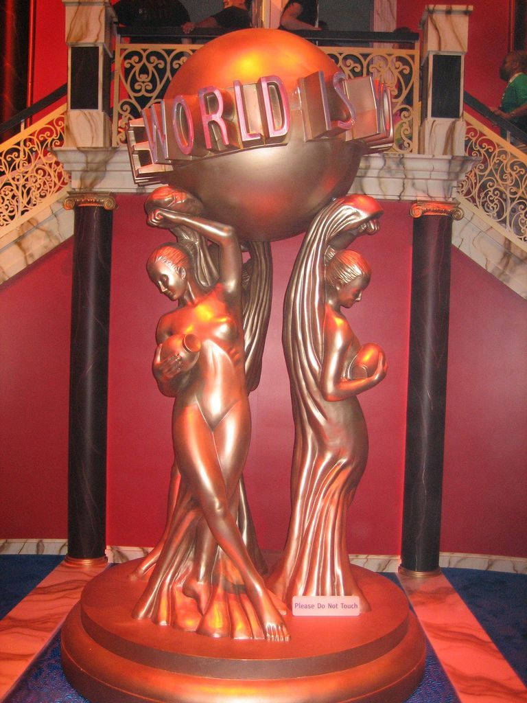 The World Is Yours 桌布 壁紙 — 兩座銅像支撐著一個帶有「WORLD」字樣的地球儀，位於裝飾華麗的劇院大廳，配有紅色牆壁和大理石柱。