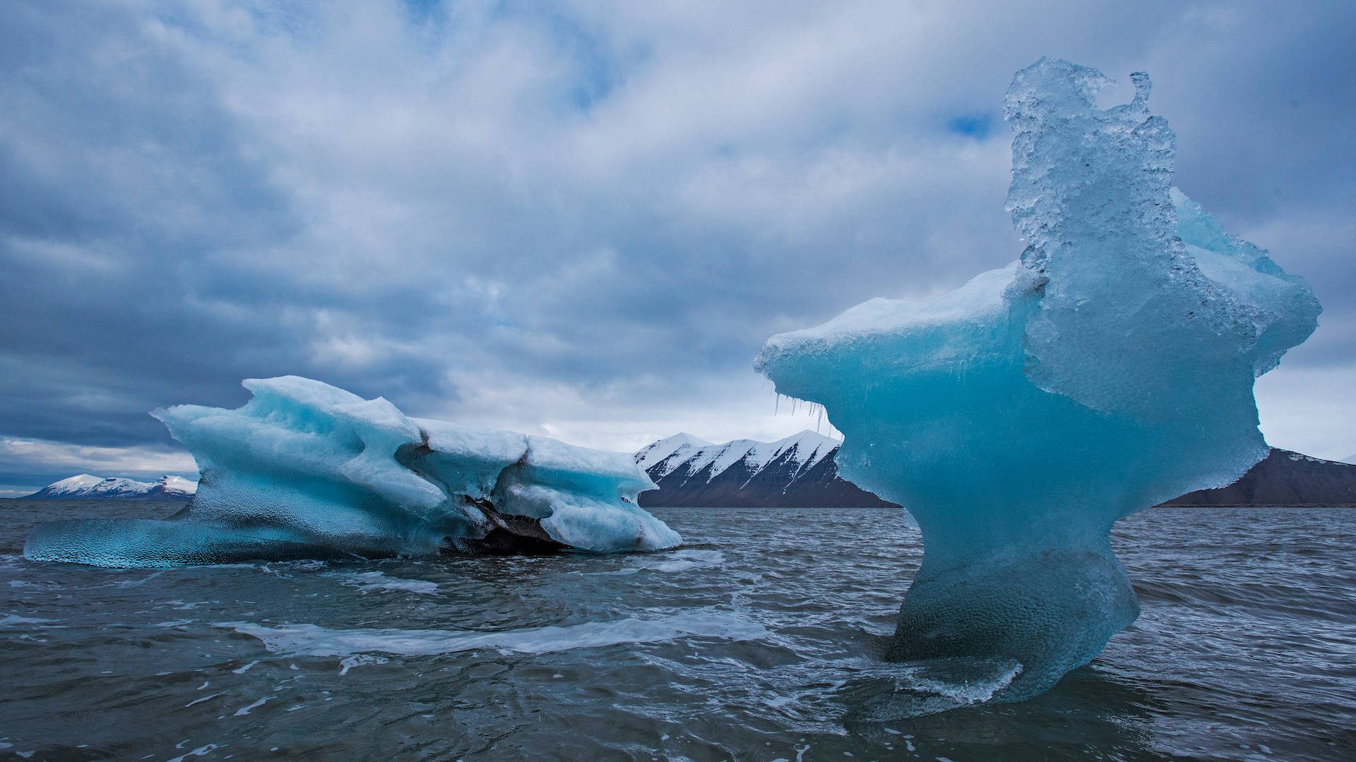 Svalbard 桌布 壁紙 — 兩座雄偉的藍綠色冰山在波濤洶湧的水域中漂浮，遠處是白雪覆蓋的山脈，天空多雲而富有戲劇性。