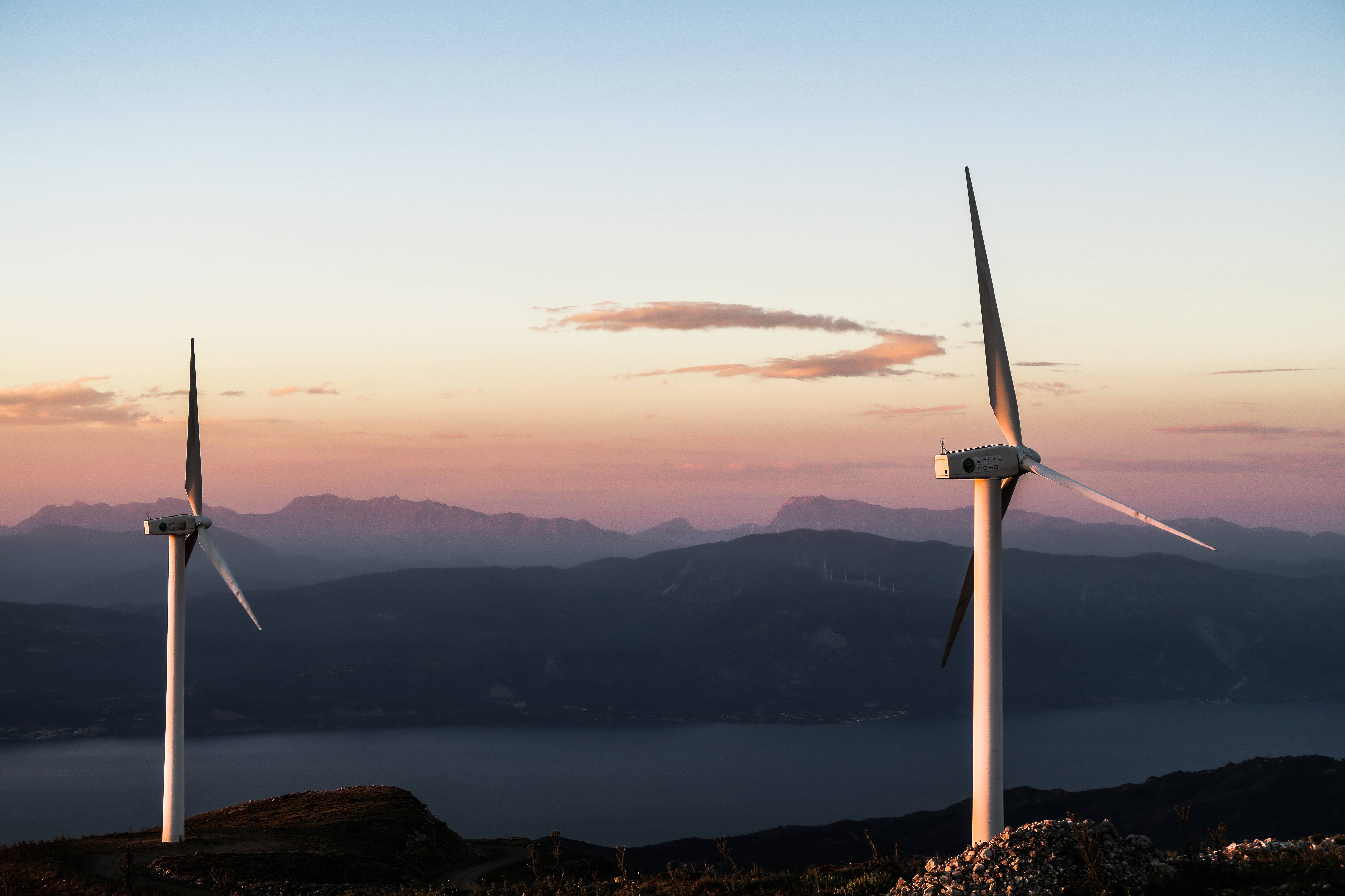 Energy 桌布 壁紙 — 兩座風力發電機聳立在山坡上，夕陽西下，在粉彩色的天空襯托下形成剪影，遠方有山巒，下方是平靜的海面。