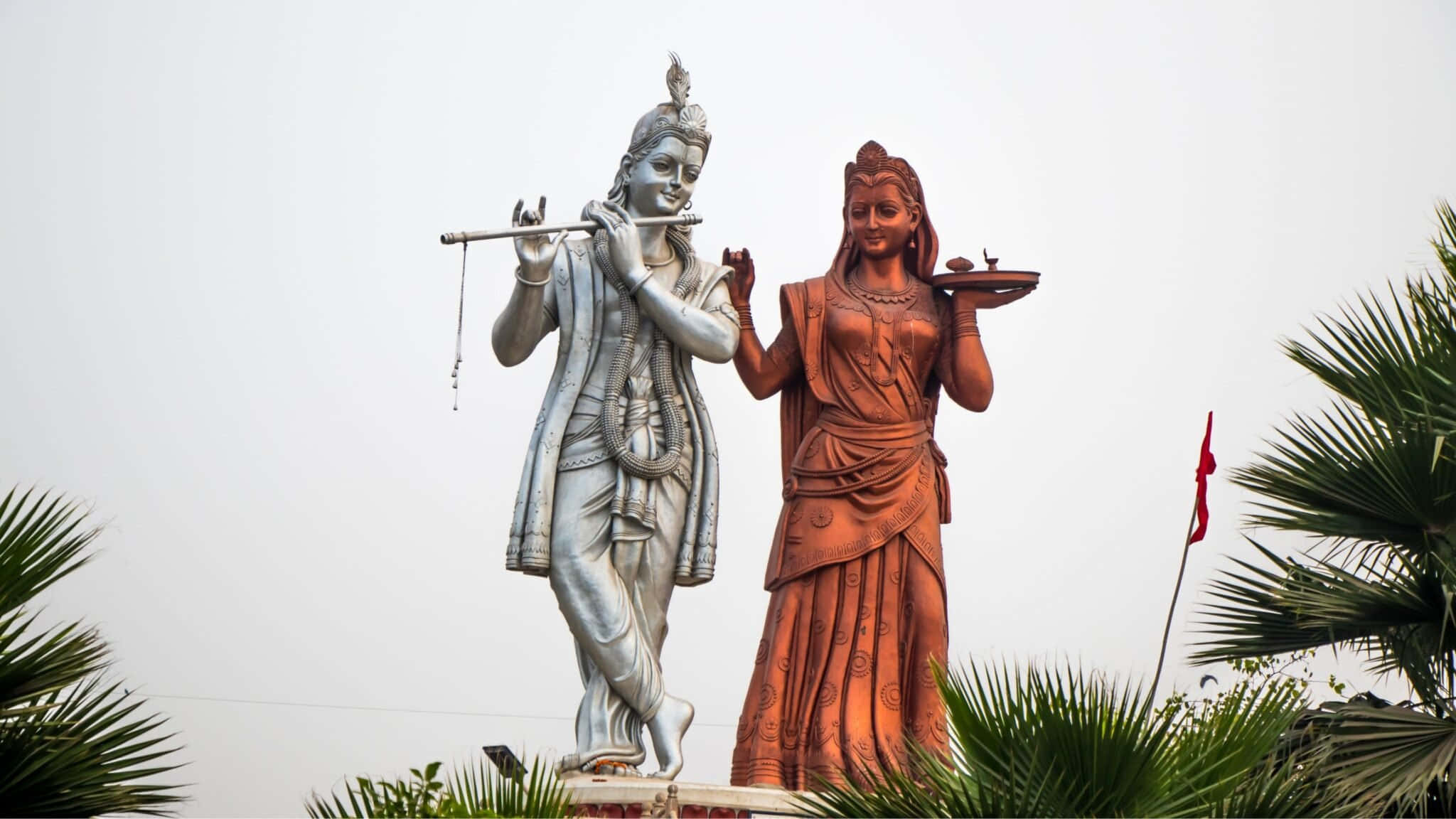 statue 桌布 壁紙 — 兩座高聳的雕像，一位銀色吹笛者和一位青銅獻祭者，站在棕櫚葉中，天空多雲。