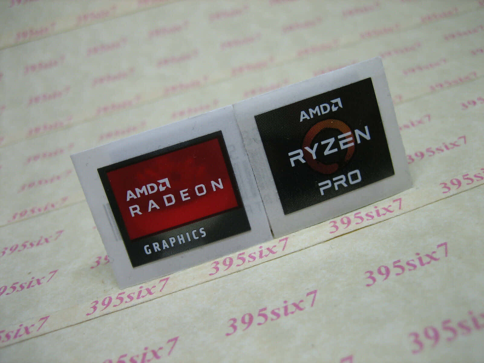 AMD 桌布 壁紙 — 兩張AMD處理器貼紙（Radeon Graphics和Ryzen Pro）放在帶有重複395six7文字的花紋表面上。