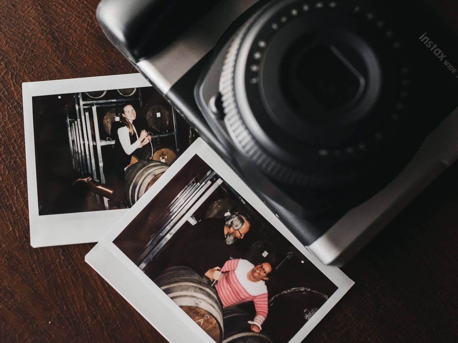 Photograph 桌布 壁紙 — 兩張即時照片，顯示酒窖中的人們，旁邊放著一部黑色Instax相機，置於深色木質表面上。