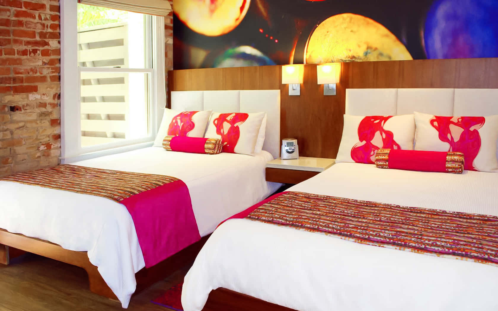 Hotel Room 桌布 壁紙 — 兩張單人床配有白色床單和鮮紅色枕頭，位於磚牆和窗戶旁邊的木製床頭板上的宇宙壁畫下方。