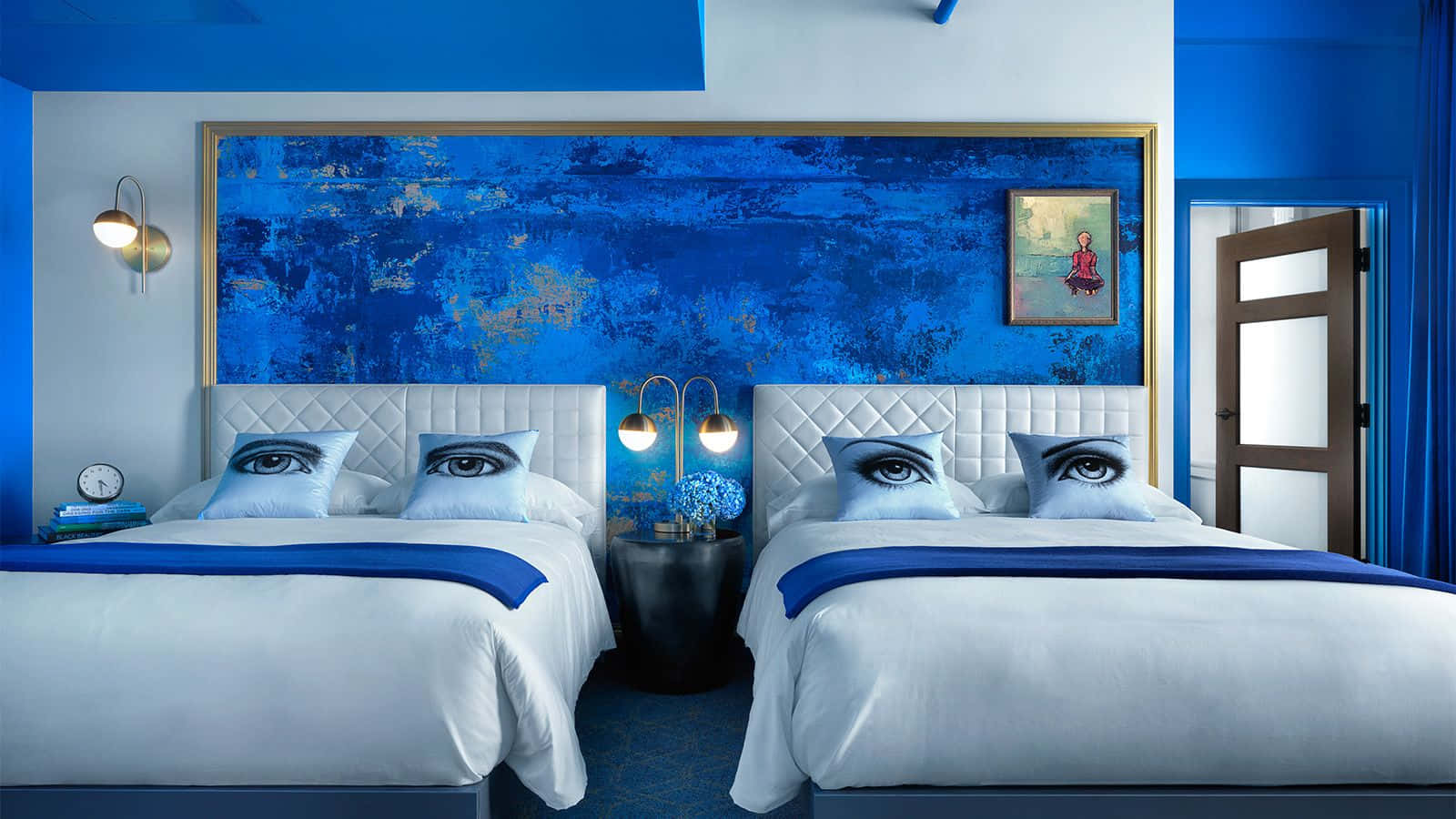 Hotel Room 桌布 壁紙 — 兩張對稱的床，配有眼睛圖案的枕頭，面向現代酒店房間中大膽的藍色紋理牆壁藝術。