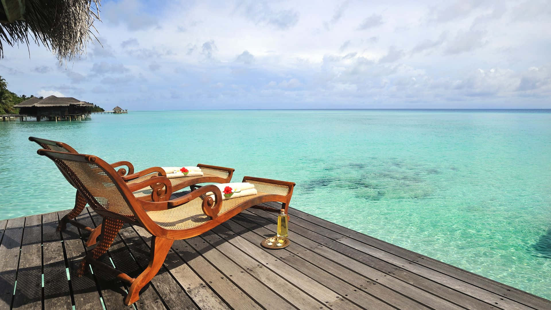 Maldives Beach 桌布 壁紙 — 兩張帶有紅玫瑰點綴的木製躺椅，放置在一個飽經風霜的甲板上，俯瞰著在部分多雲天空下的碧綠海水。