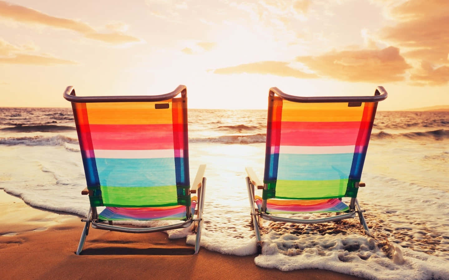 Beach Sunbed 桌布 壁紙 — 兩張彩虹條紋沙灘椅面向海洋，金色夕陽下，溫柔的海浪拍打著海岸。