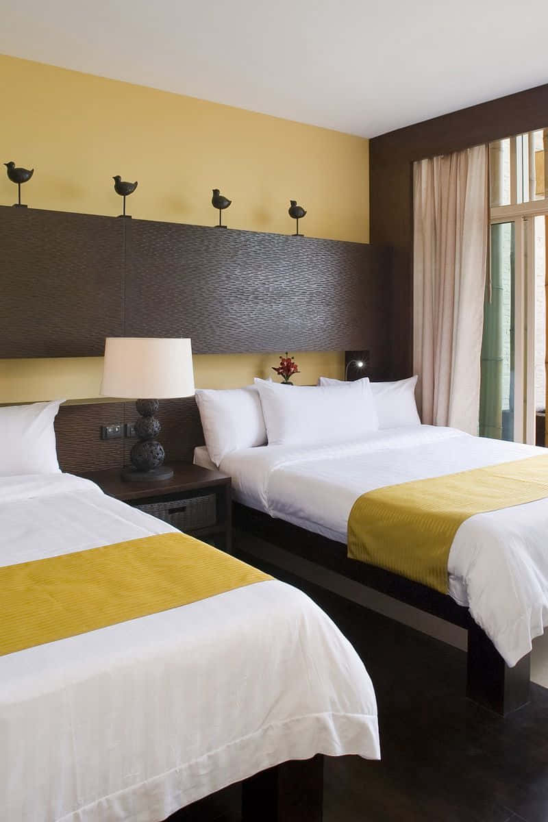 Hotel Room 桌布 壁紙 — 兩張整齊鋪好的床，配有白色床單和黃色點綴，位於帶有鳥類雕像裝飾的紋理床頭板下方。