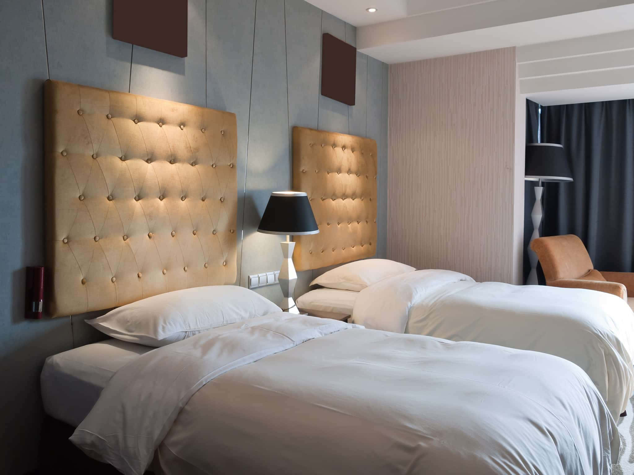 Hotel Room 桌布 壁紙 — 兩張整齊鋪好的床，配有簇絨米色床頭板，在現代飯店房間的環境照明下溫暖地發光。