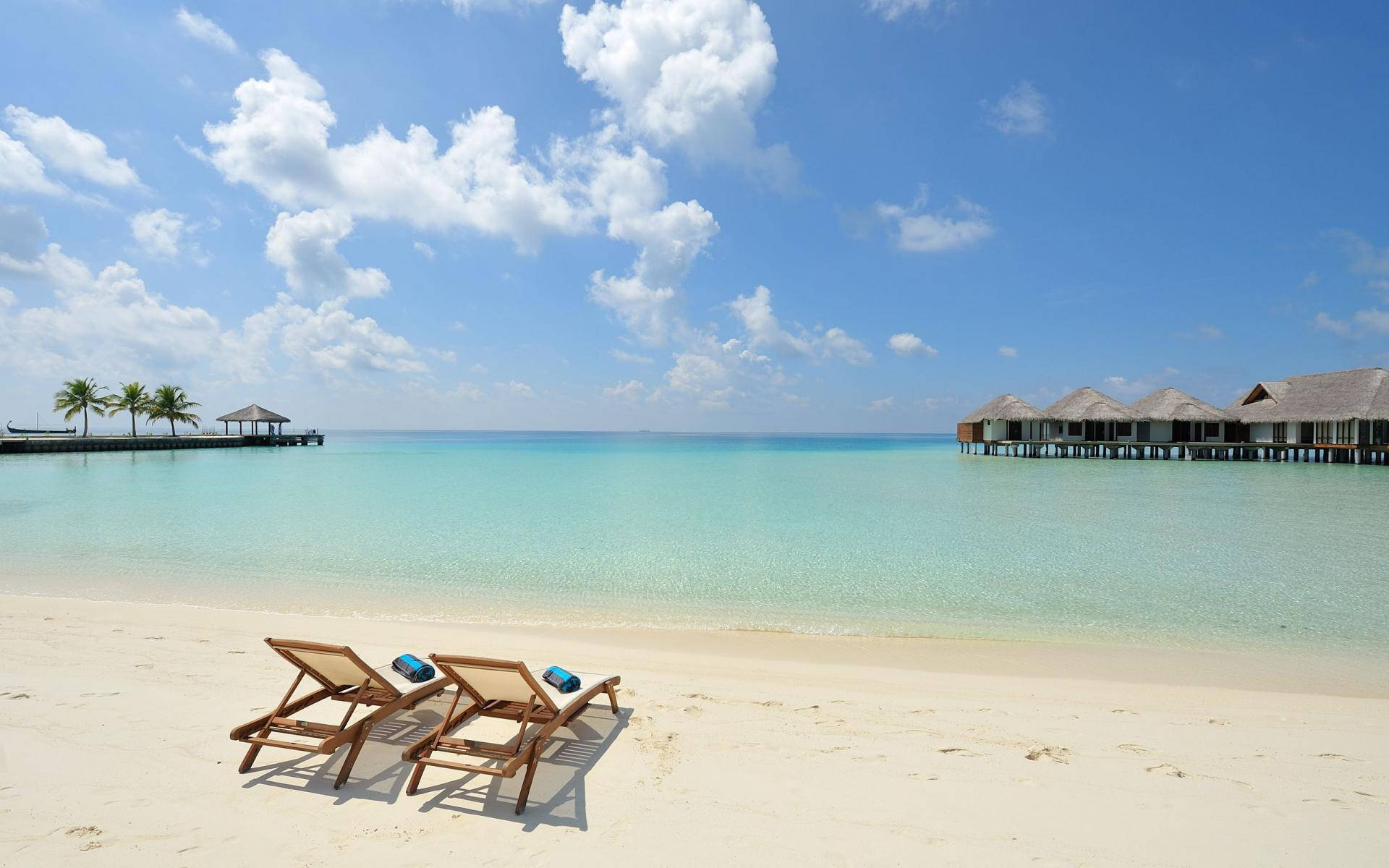 Maldives 桌布 壁紙 — 兩張木製躺椅放在純白沙灘上，面向碧綠海水和水上平房，天空湛藍，白雲朵朵。