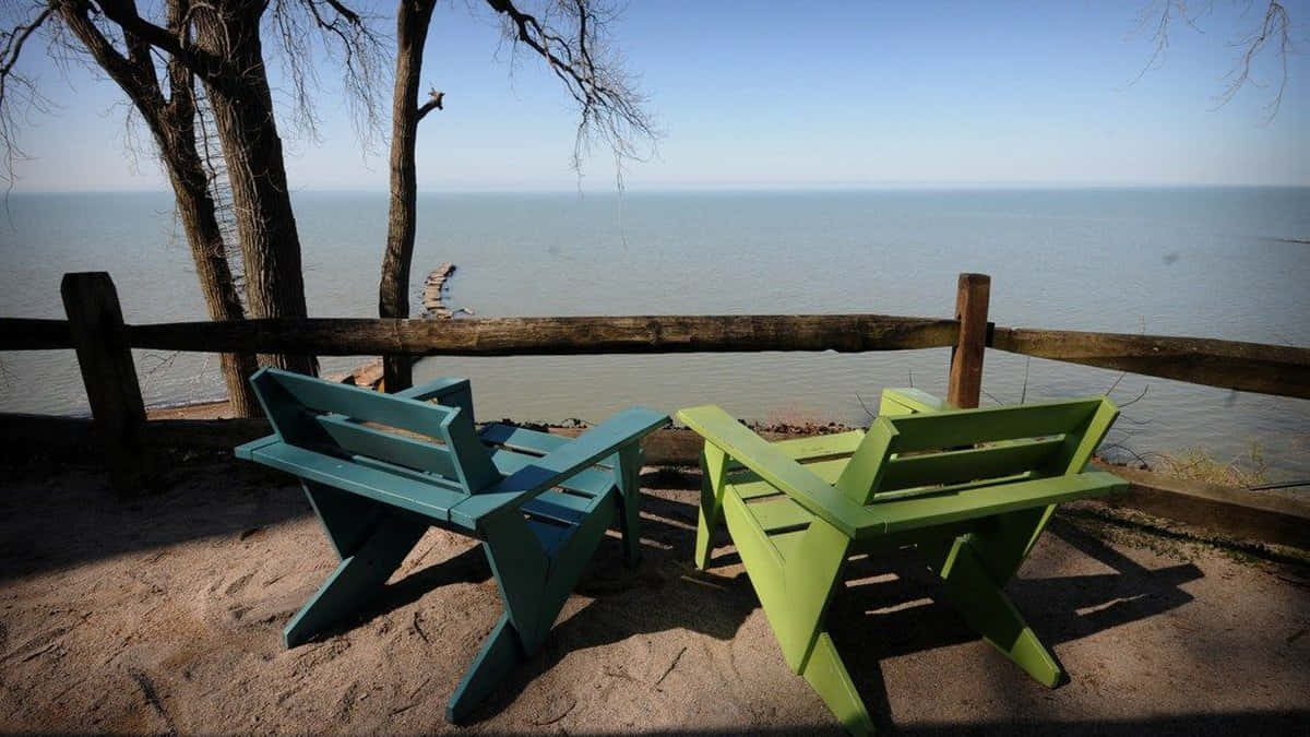 Lake Erie 桌布 壁紙 — 兩張色彩繽紛的木椅面向平靜的湖泊，在清澈的藍天下，光禿的樹木投下陰影。