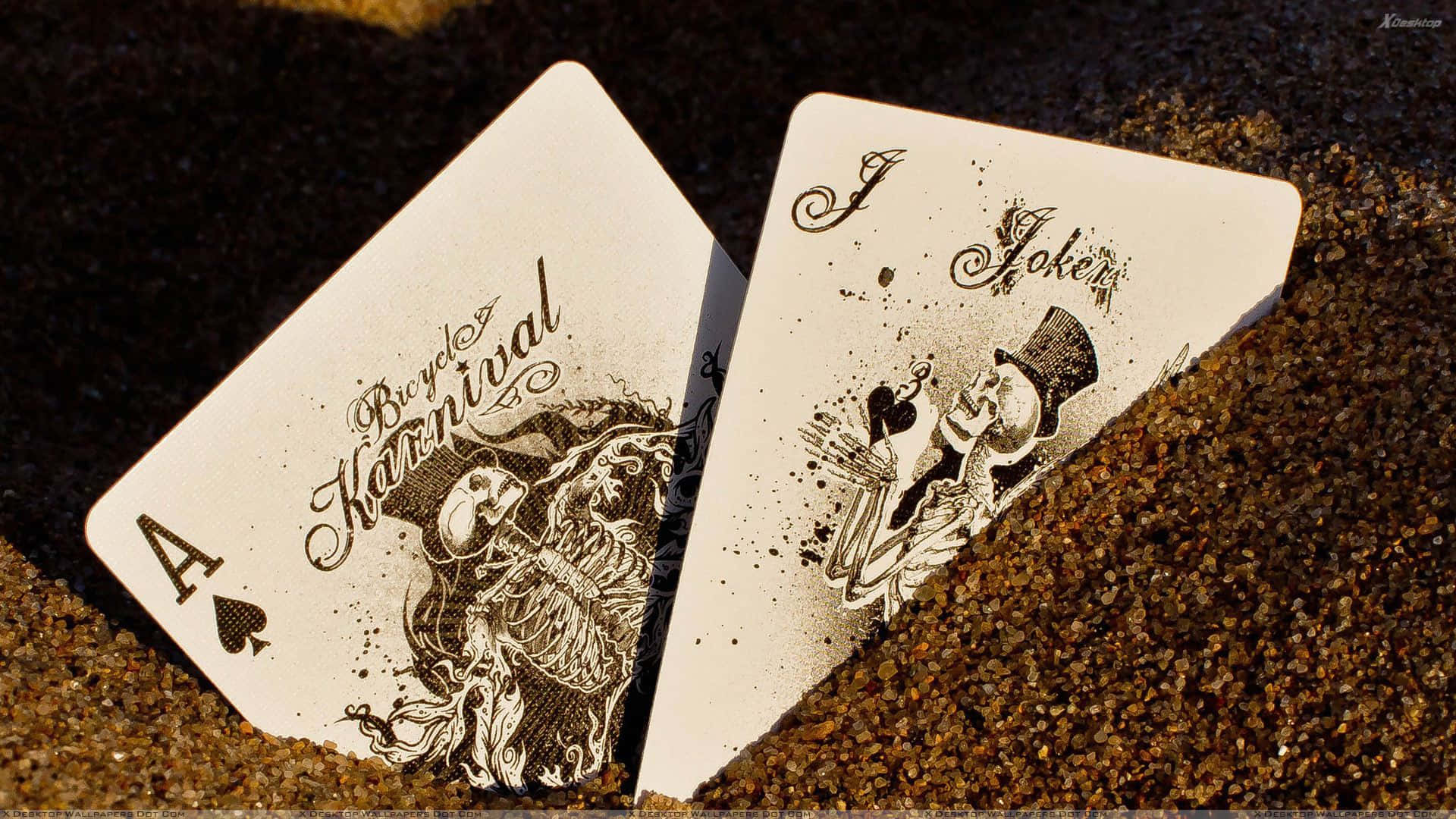 Poker 桌布 壁紙 — 兩張華麗的撲克牌，一張黑桃A和一張小丑牌，躺在溫暖陽光下的粗糙沙地上。