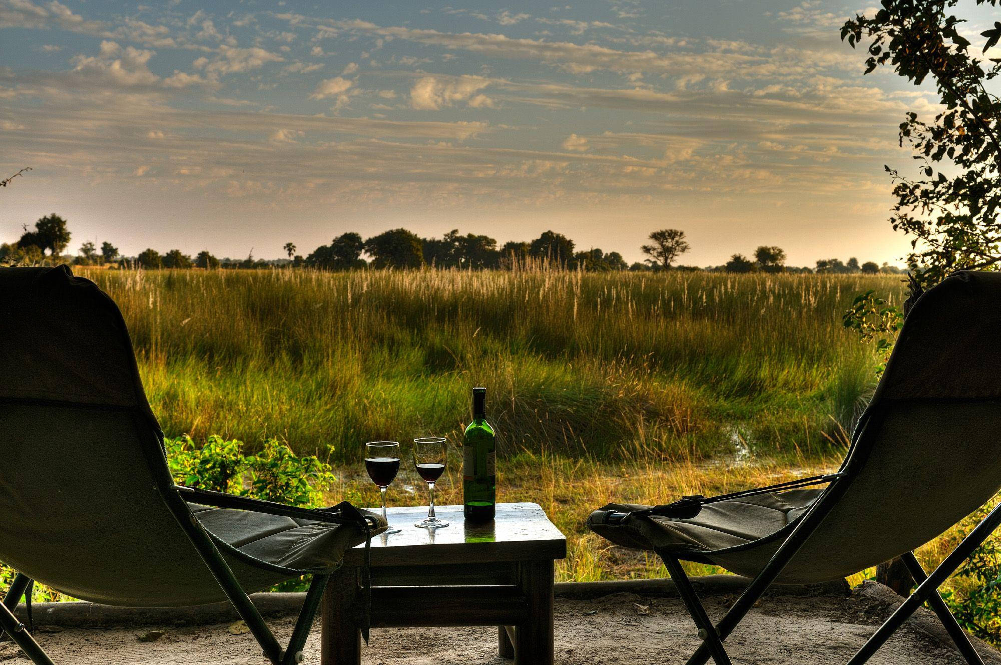 Botswana 桌布 壁紙 — 兩張露營椅面向日落時分的金色草地，小桌子上有酒瓶和酒杯。