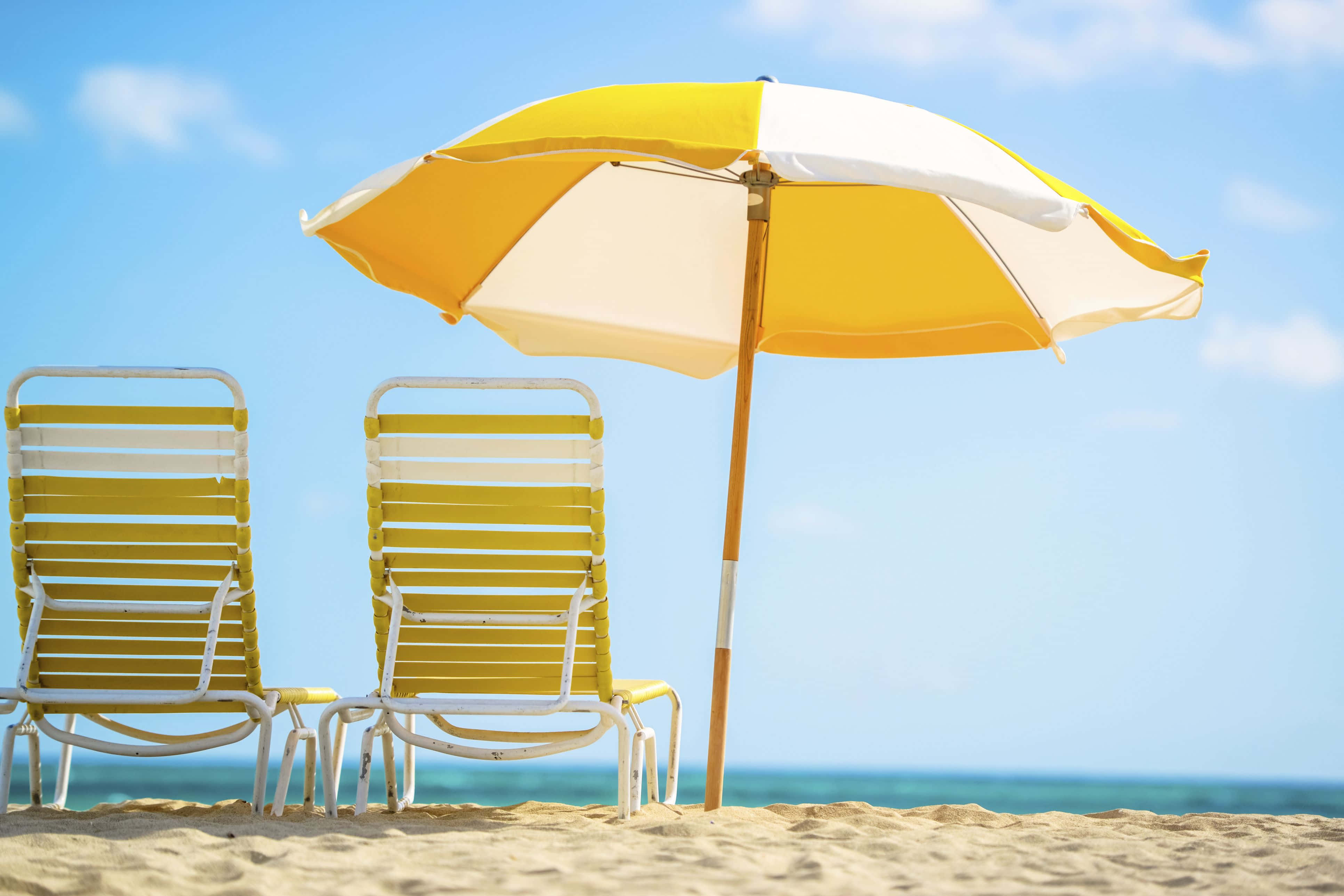 Beach Umbrella 桌布 壁紙 — 兩張黃色沙灘椅放在黃白相間的遮陽傘下，位於金色沙灘上，面向碧綠的海洋，天空清澈湛藍。
