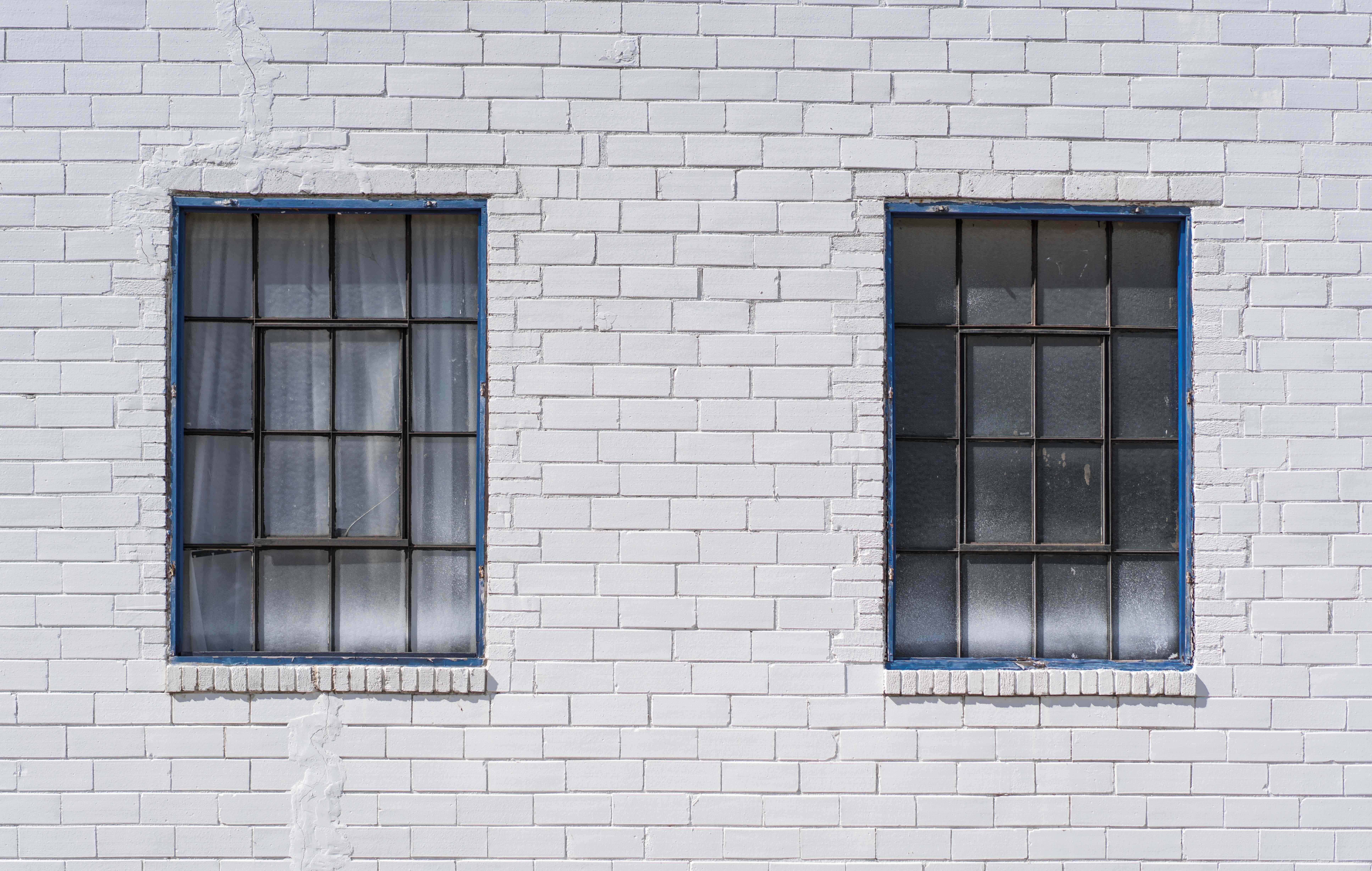 White Brick Wall 桌布 壁紙 — 兩扇經風化的藍框窗戶帶有格柵條，並排立在白色磚牆上，處於明亮日光下。