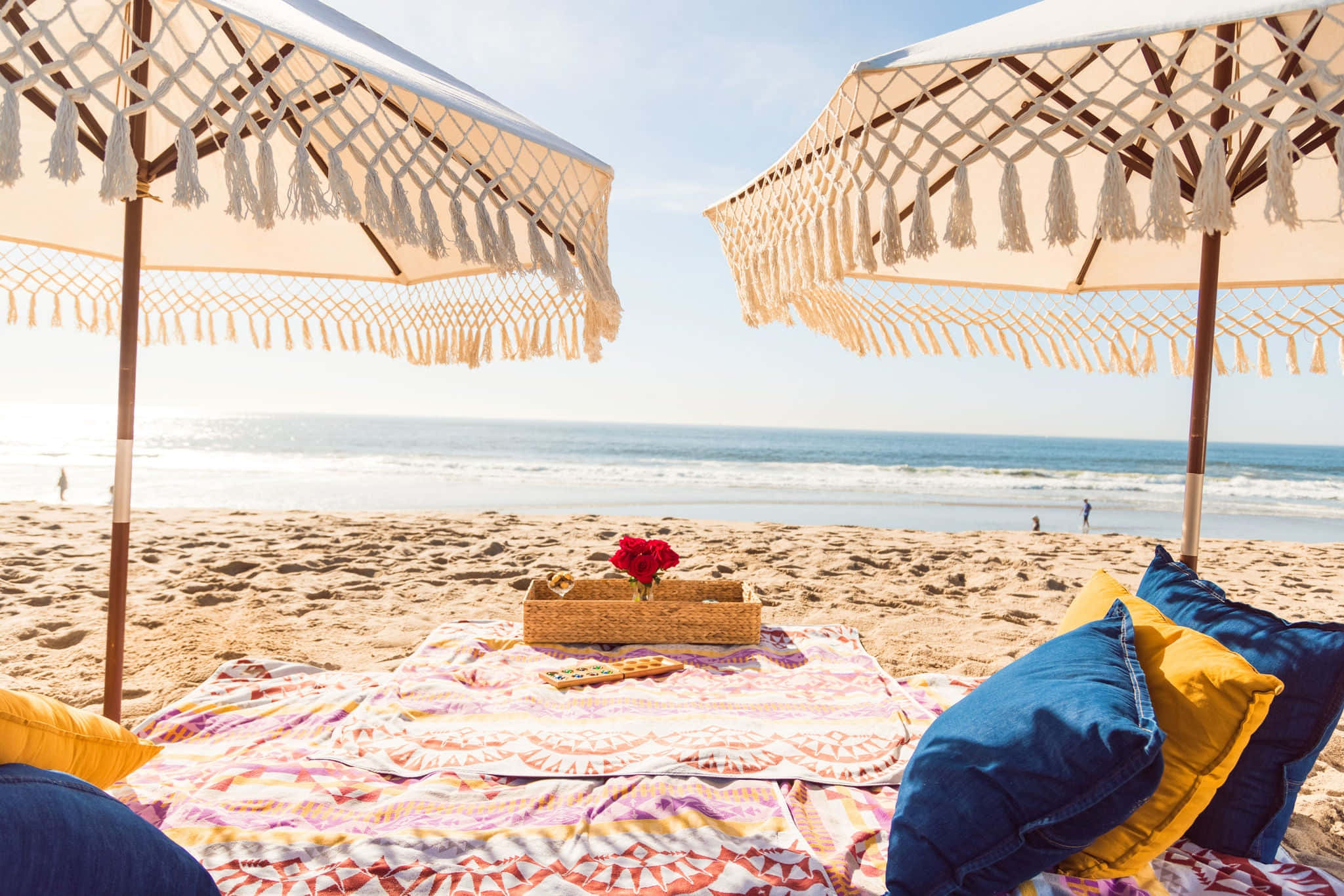 Beach Picnic 桌布 壁紙 — 兩把macrame沙灘傘為一個色彩繽紛的毯子（配有枕頭和一朵紅花）在陽光明媚的沙灘上遮蔭。