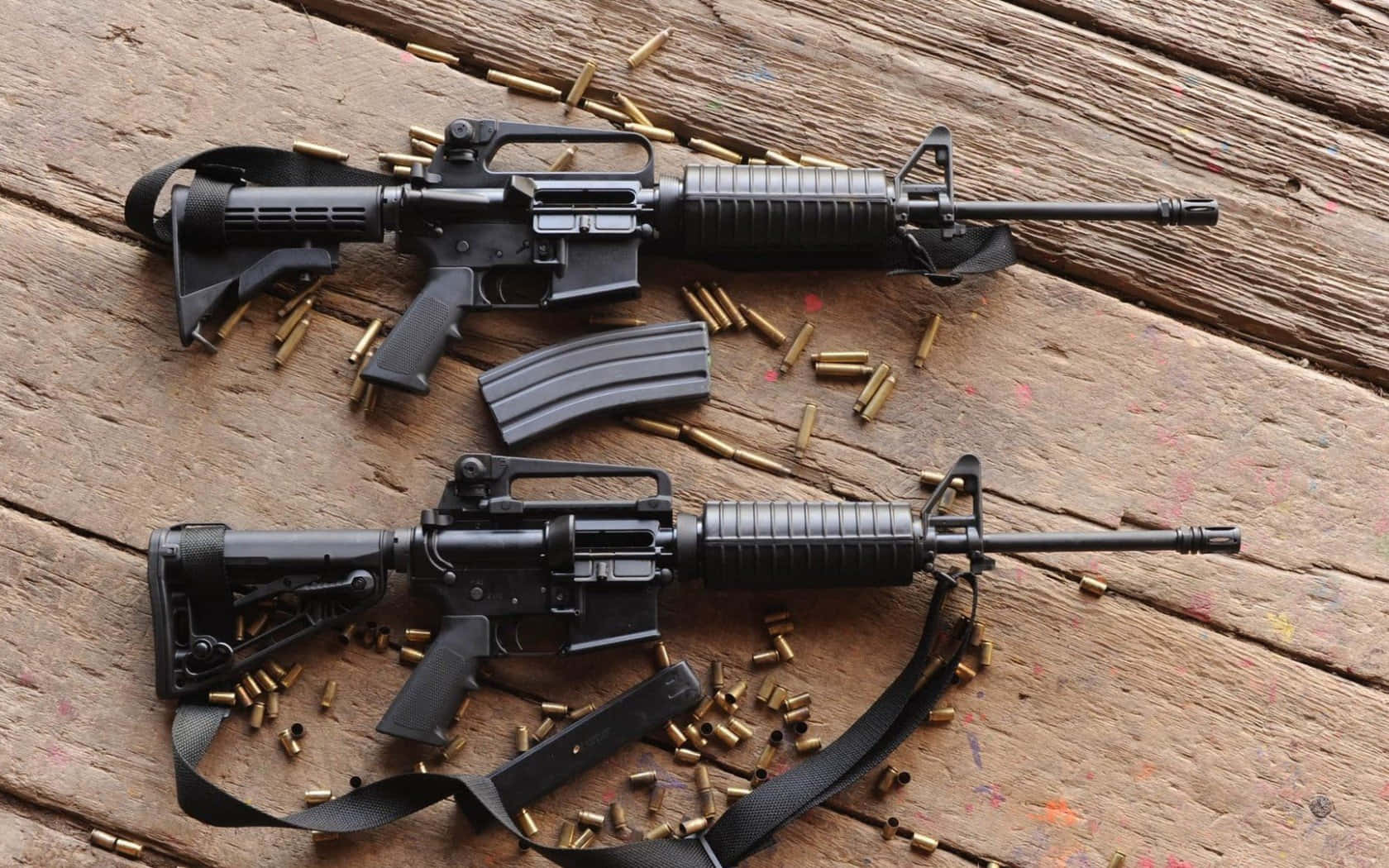 Rifle 桌布 壁紙 — 兩把黑色突擊步槍放在經風化的木質表面上，周圍散落著黃銅彈殼。