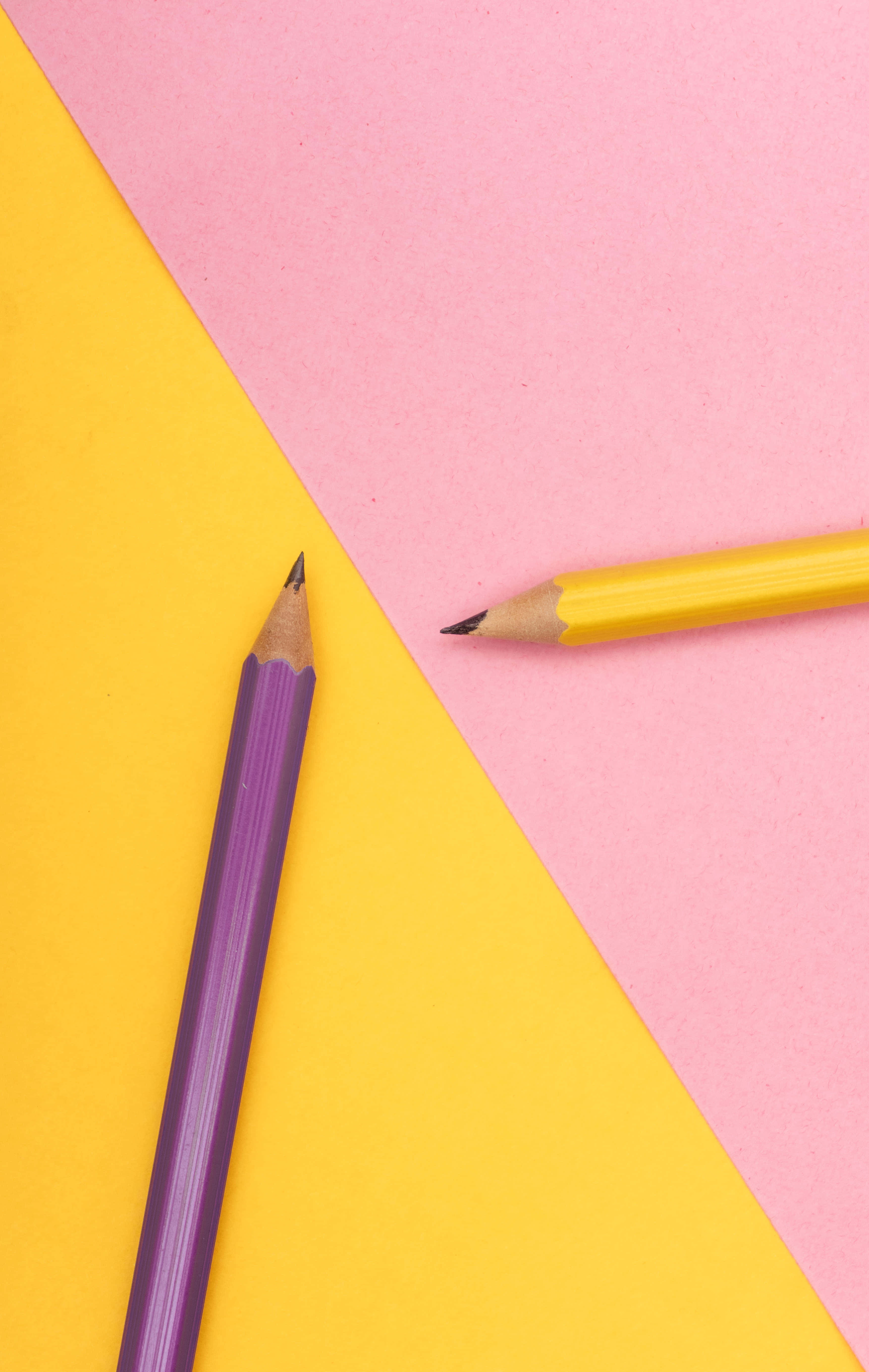 pencil 桌布 壁紙 — 兩支削尖的彩色鉛筆斜放在充滿活力的黃色和粉紅色幾何背景上。