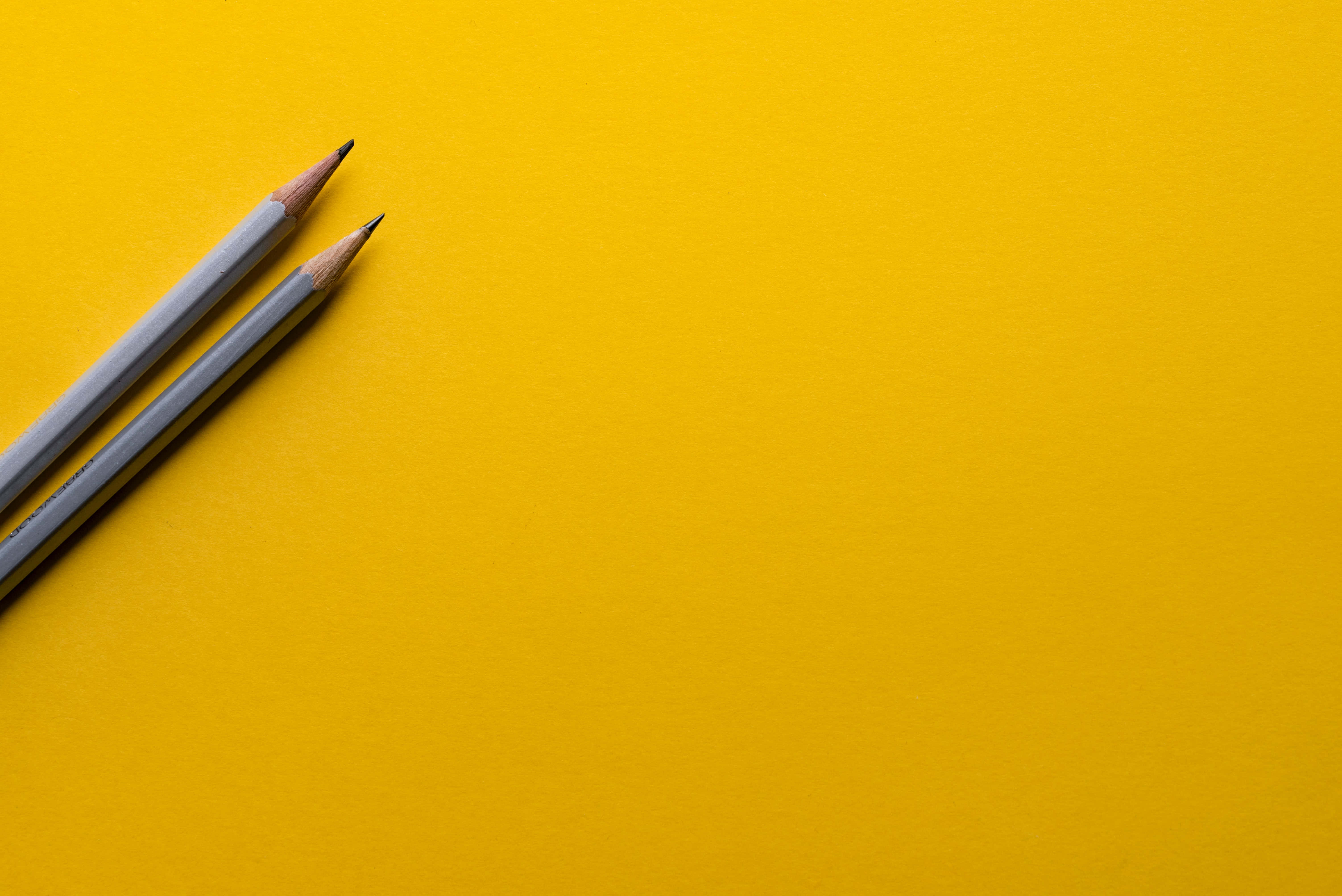 pencil 桌布 壁紙 — 兩支削尖的灰色鉛筆對角放置在充滿活力的黃色背景上，留有充足的負空間。