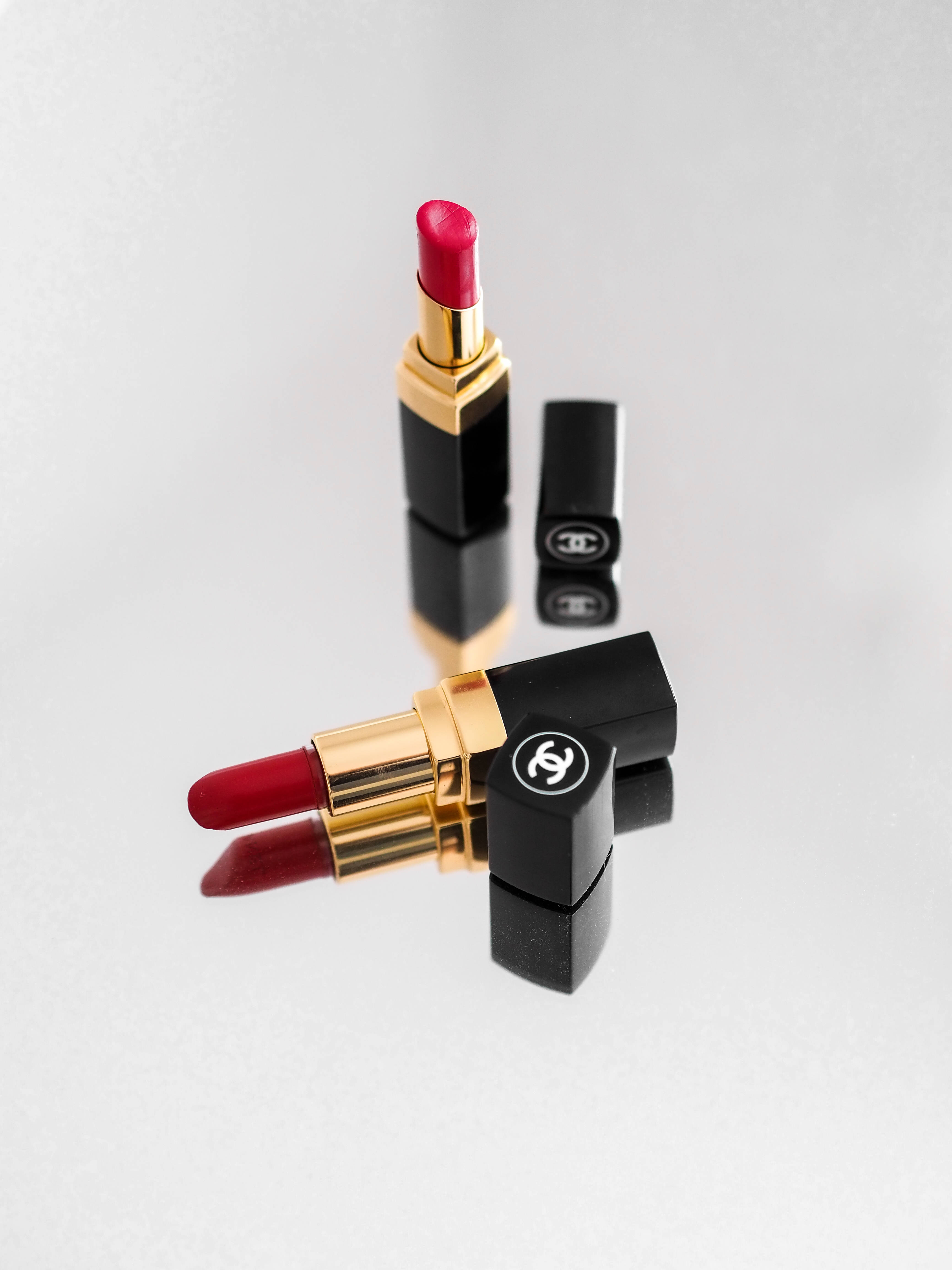Chanel 桌布 壁紙 — 兩支帶有鮮豔紅色色素和金色點綴的Chanel唇膏，優雅地放置在反光的白色表面上。