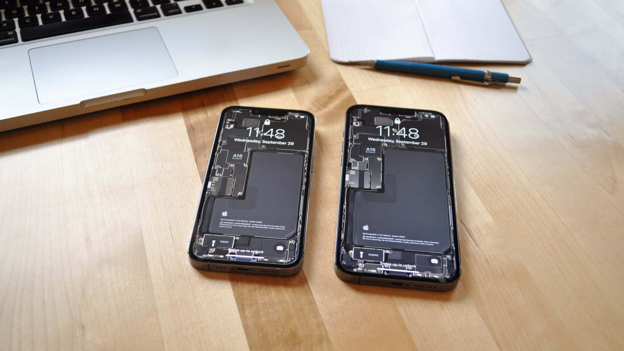 Smartphone 桌布 壁紙 — 兩支拆解的智慧型手機，內部零件外露，放在木桌上，旁邊有一台筆記型電腦和一本筆記本。