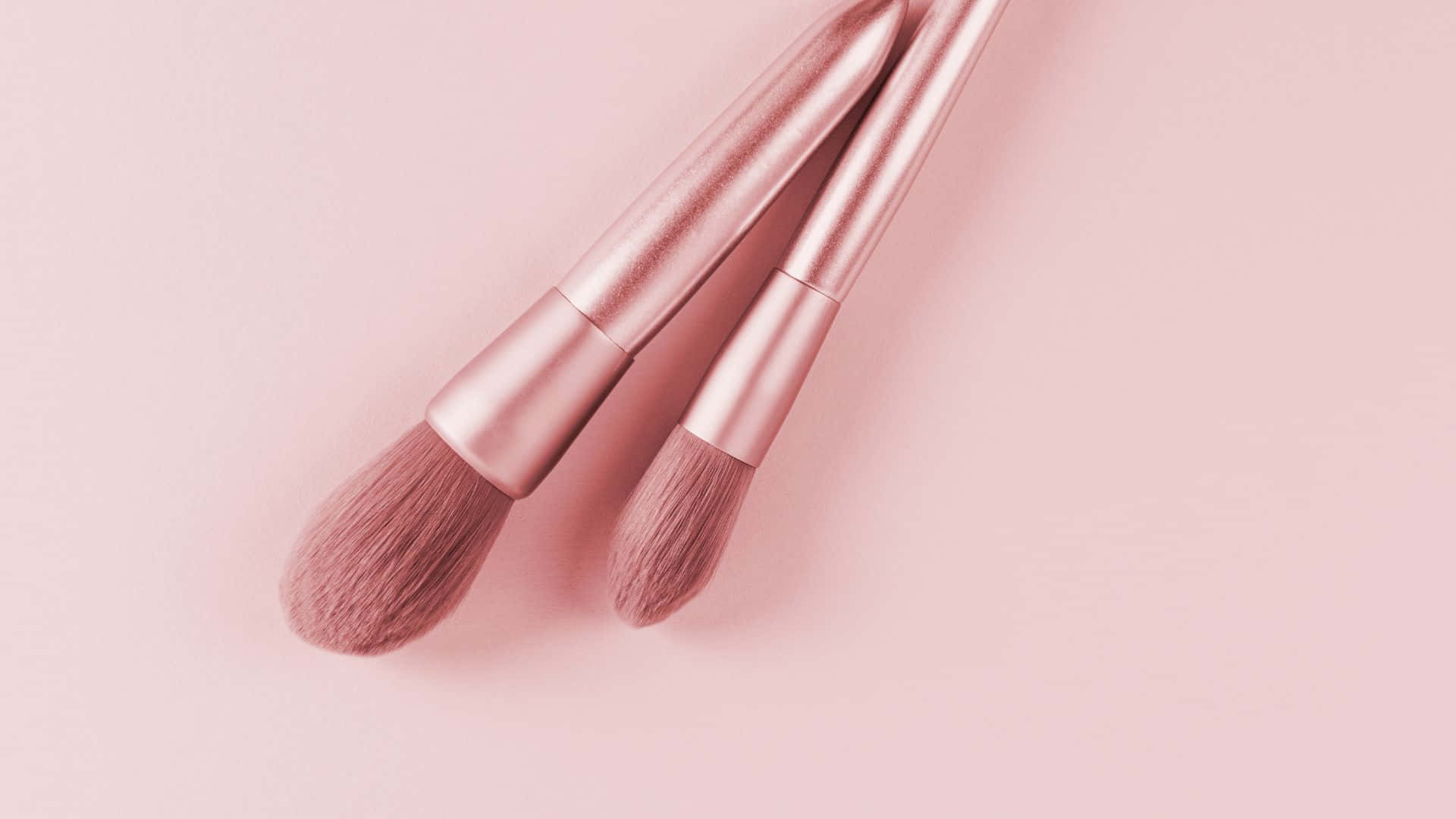 Pink Makeup 桌布 壁紙 — 兩支玫瑰金化妝刷斜放在柔和的粉紅色背景上，帶有輕柔的陰影。