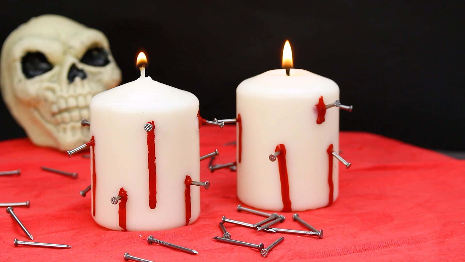 Halloween Candles 桌布 壁紙 — 兩支白色蠟燭帶有釘子和紅色蠟滴，在黑色背景的紅布上，於一個頭骨旁邊燃燒。