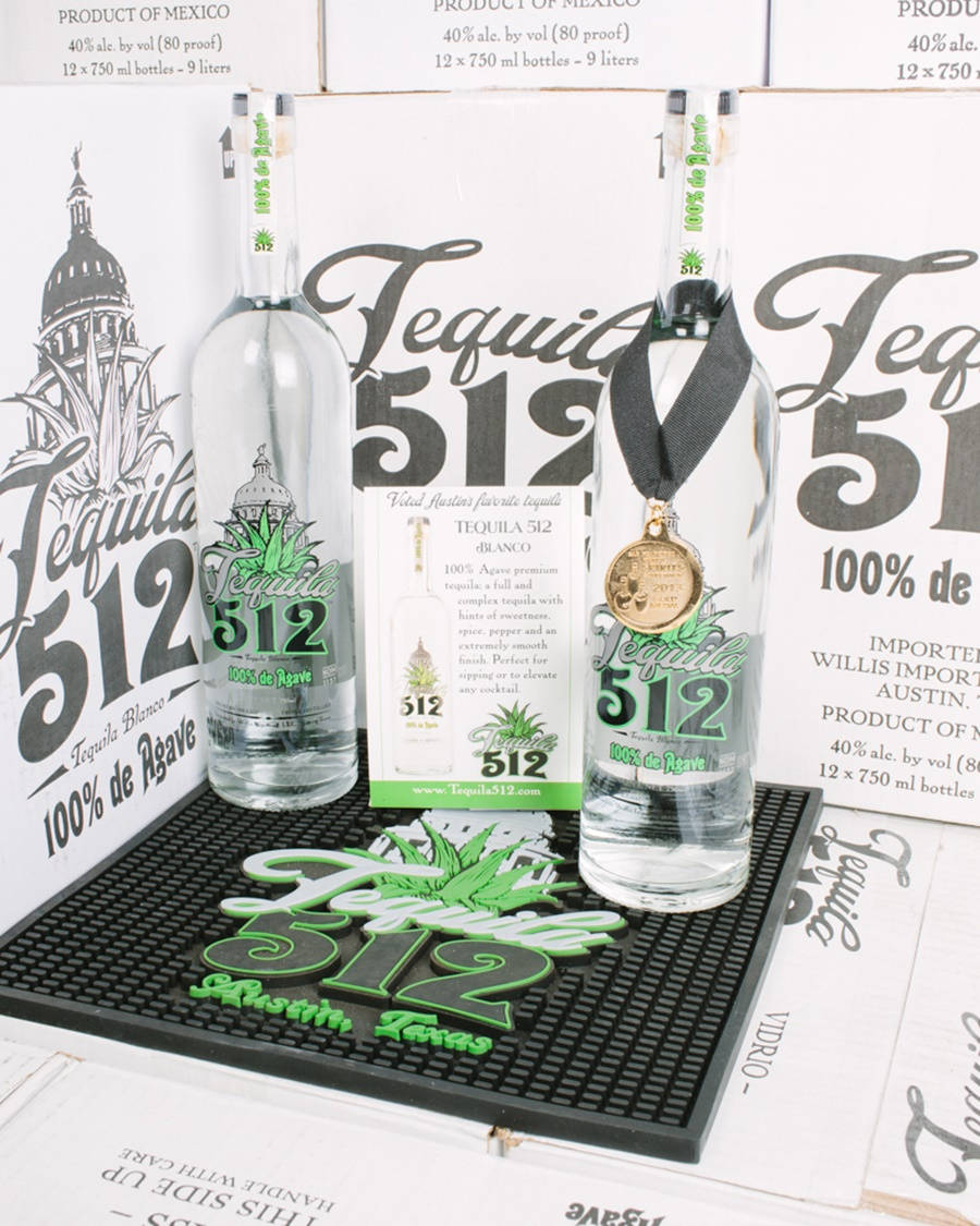 drinks 桌布 壁紙 — 兩支透明龍舌蘭酒瓶，帶有綠色標籤和一枚金牌，放在品牌墊上，背景是堆疊的產品盒。
