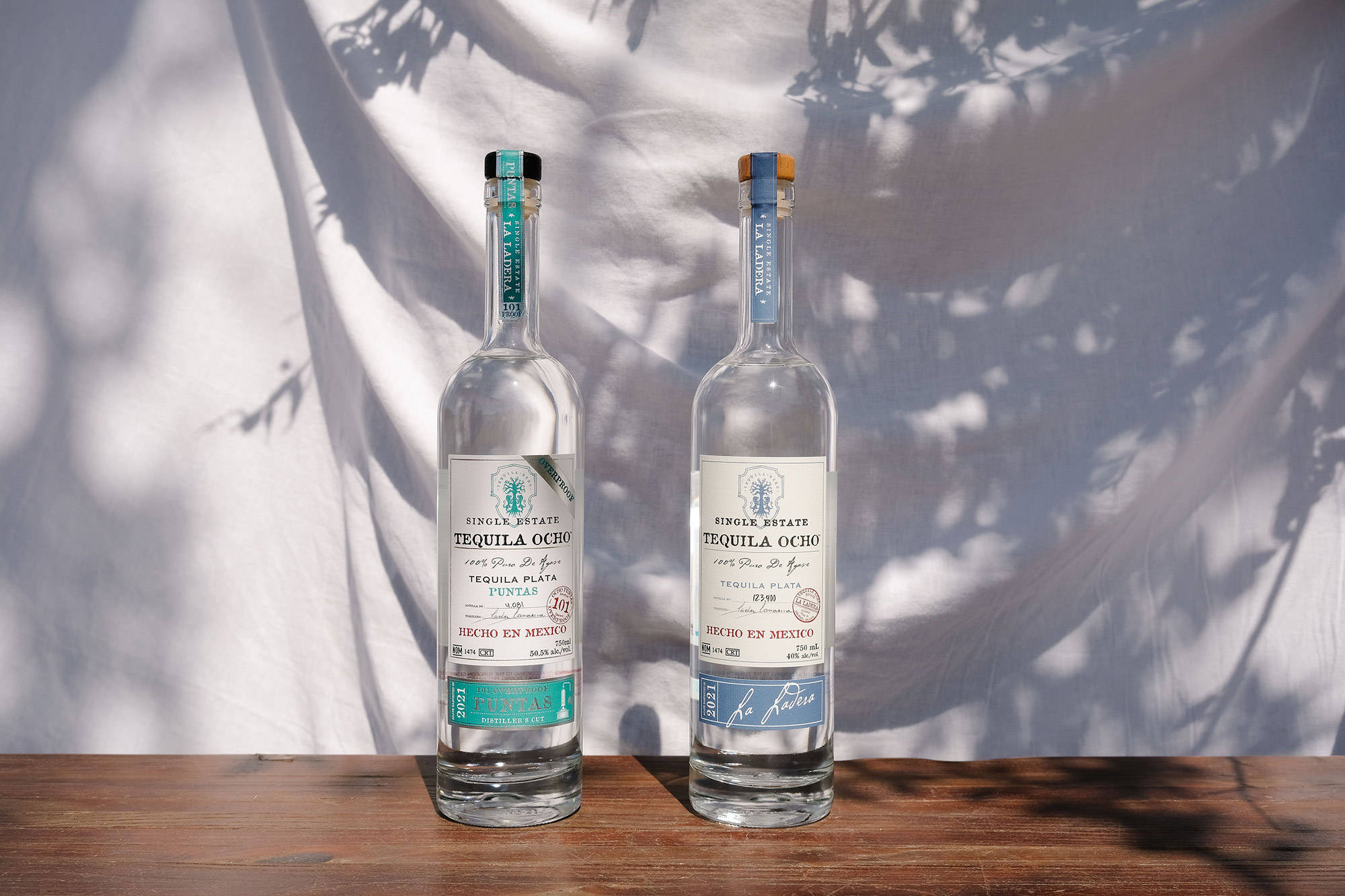 bottle 桌布 壁紙 — 兩支透明龍舌蘭酒瓶標有'Tequila Ocho'，放在木質表面上，背景是帶有樹影的白色布料。