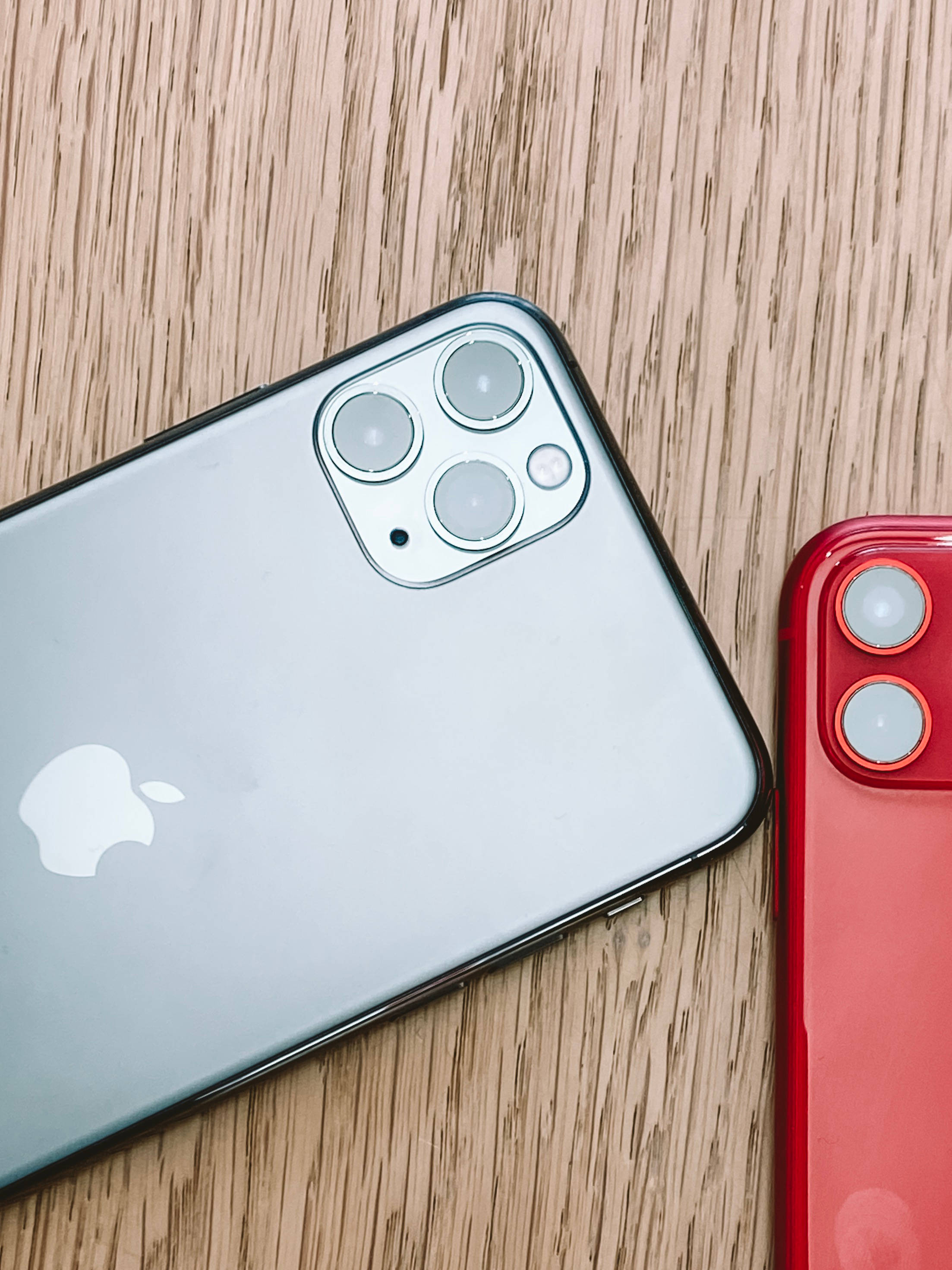 iPhone 11 桌布 壁紙 — 兩支銀色和紅色的iPhone對角線放置在溫暖的木質表面上，展示它們的相機系統。