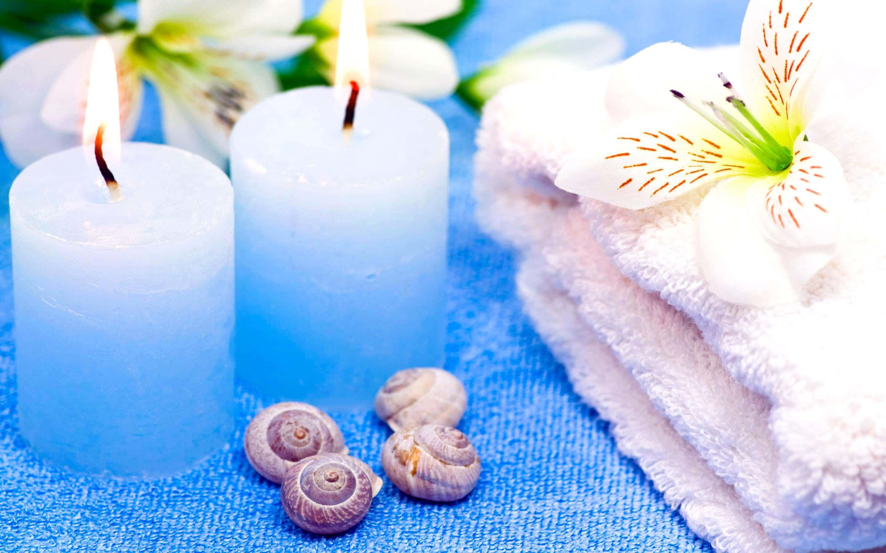 Relaxation 桌布 壁紙 — 兩支點燃的藍色蠟燭在摺疊的白色毛巾和貝殼旁邊發光，置於帶有白色花朵的紋理藍色表面上。