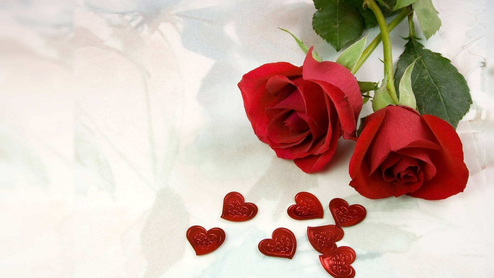 Red Roses 桌布 壁紙 — 兩朵鮮豔的紅玫瑰，帶有綠色莖，放在柔軟的白色紋理表面上，旁邊散落著紅色的心形糖果。