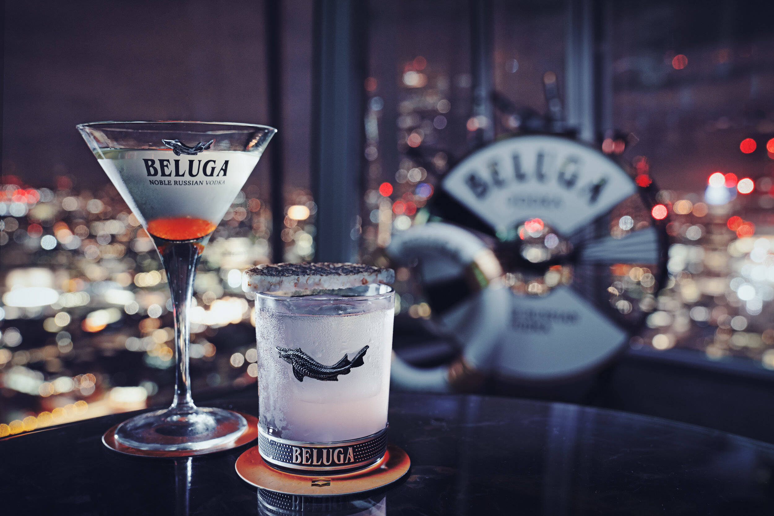 martini 桌布 壁紙 — 兩杯Beluga伏特加雞尾酒在酒吧上閃耀，背後是夜晚模糊的城市天際線發光。
