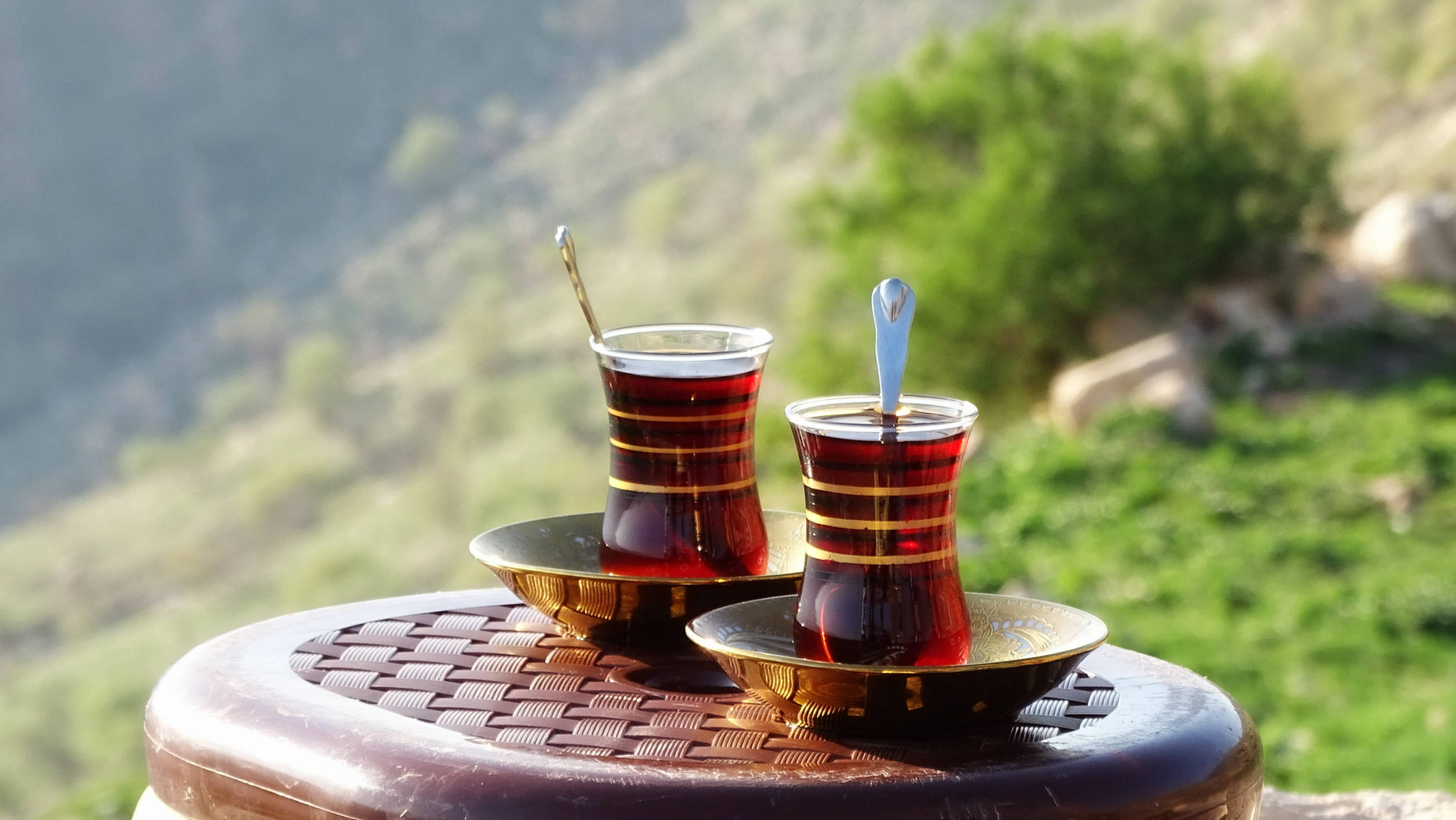 Iraq 桌布 壁紙 — 兩杯傳統土耳其茶杯連湯匙放在金色碟子上，置於編織木桌上，俯瞰陽光普照的綠色山坡。