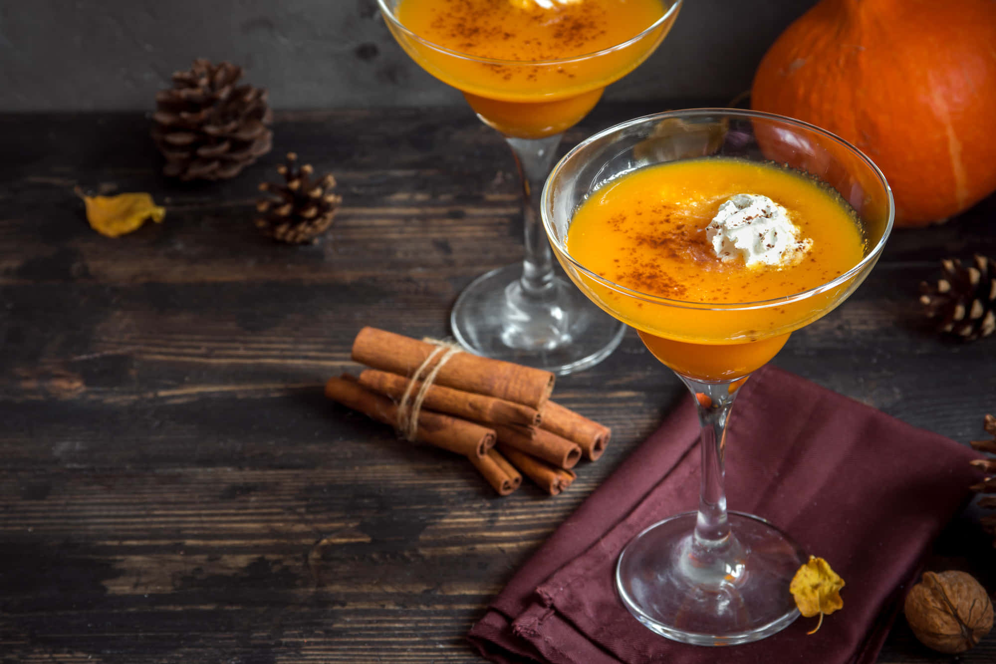 Winter Drinks 桌布 壁紙 — 兩杯充滿活力的橙色南瓜雞尾酒，飾以奶油和香料，放在質樸的木桌上，旁邊有肉桂棒、松果和一個完整的南瓜。