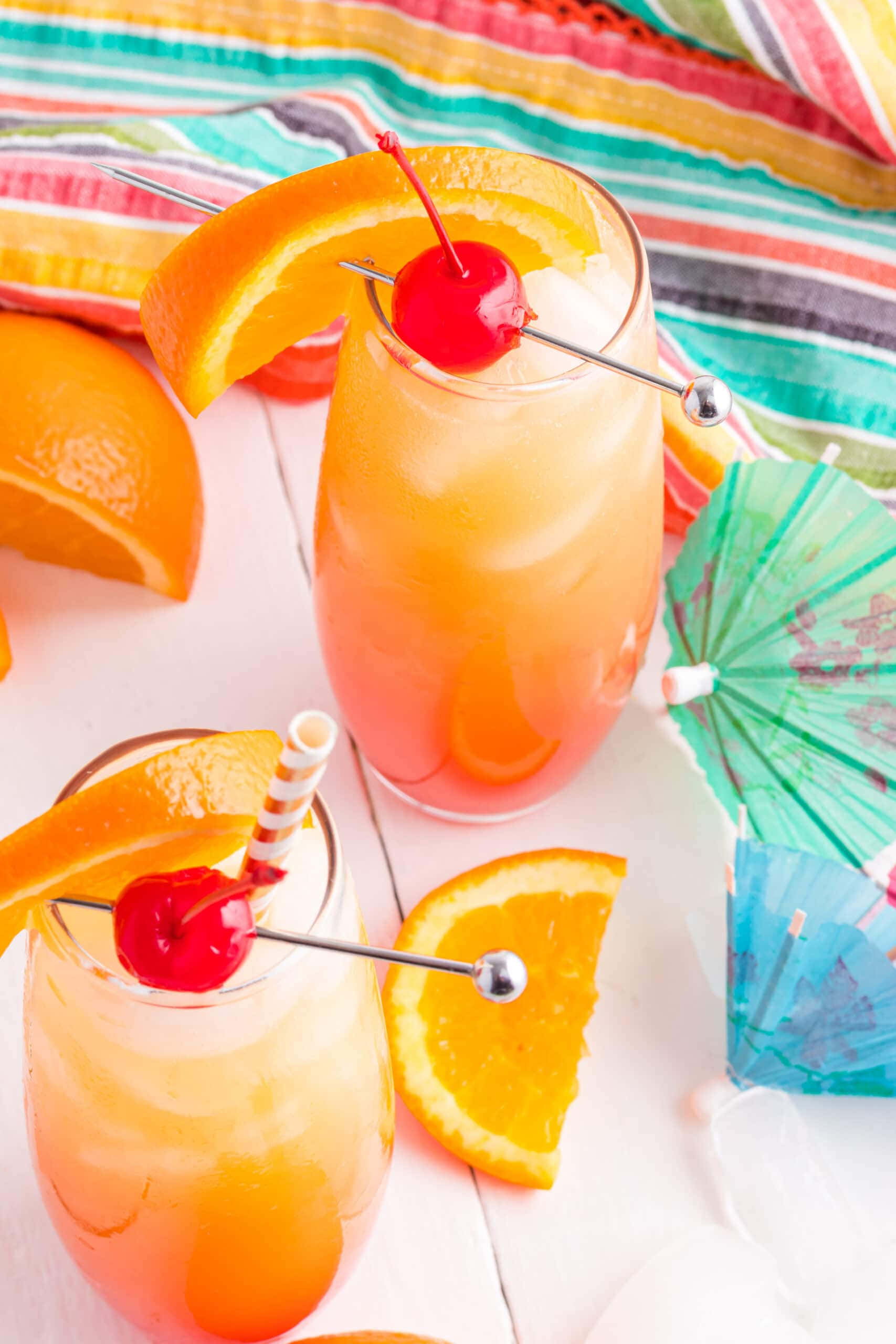 Beautiful Drink 桌布 壁紙 — 兩杯充滿活力的橙色雞尾酒，飾以櫻桃和橙片，放在色彩繽紛的條紋毛巾上，旁邊有小傘。