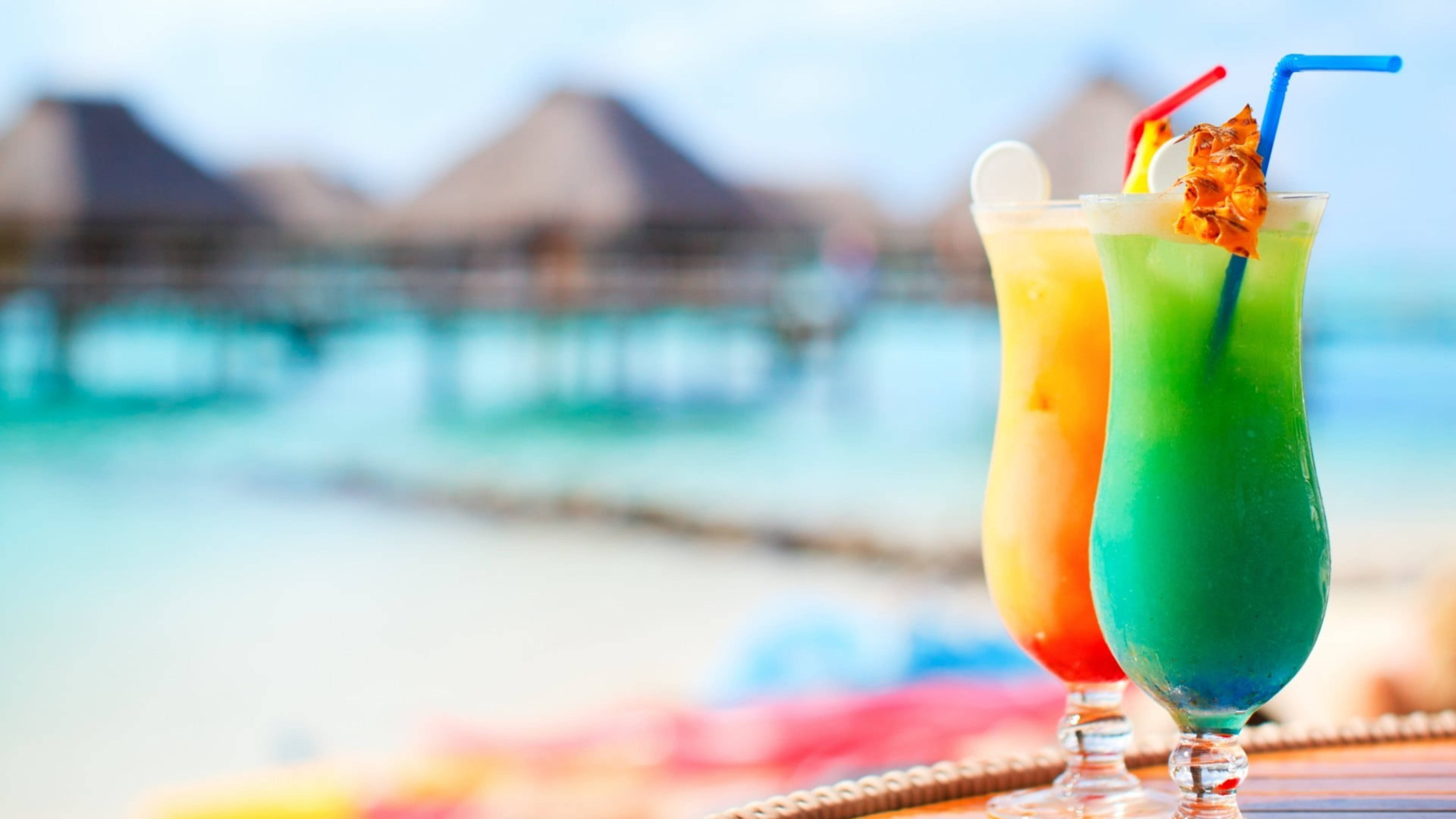 Tropical Drink 桌布 壁紙 — 兩杯充滿活力的熱帶雞尾酒，配有吸管和鳳梨裝飾，放在桌子上，背景是模糊的海灘和水上平房。