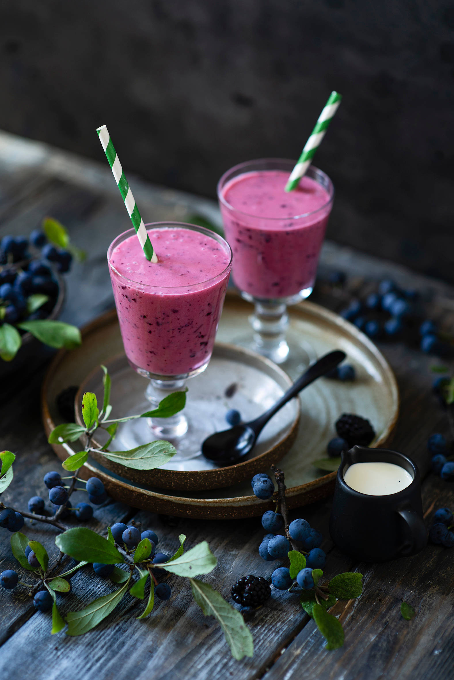 Smoothie 桌布 壁紙 — 兩杯充滿活力的粉紅藍莓冰沙放在玻璃杯中，帶有條紋吸管，放置在質樸的木桌上，周圍是新鮮漿果和綠色植物。